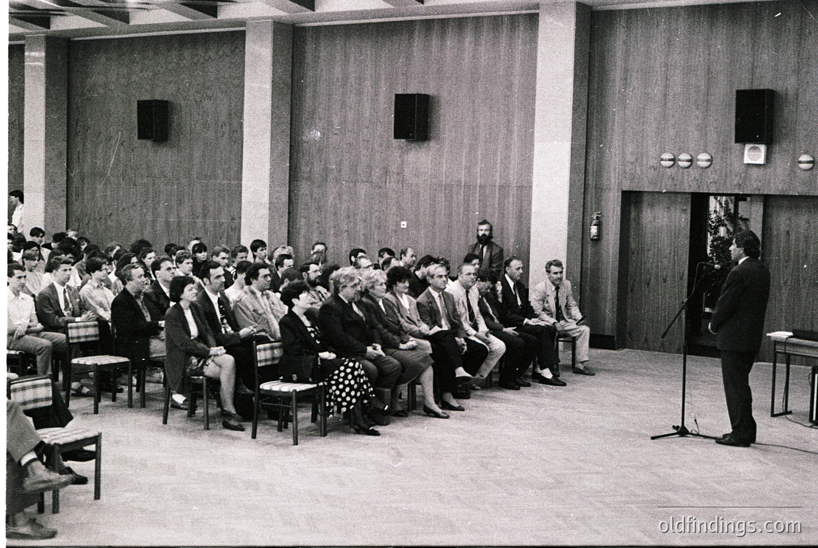 A formal indoor gathering in a mid-century conference hall, featuring a speaker at a podium addressing an attentive audience seated in rows of chairs. Wood-paneled walls and built-in speaker grilles suggest institutional or governmental use. Attire reflects 1960s–1970s office culture: suits, ties, and modest dresses. Possible educational, corporate, or political event.