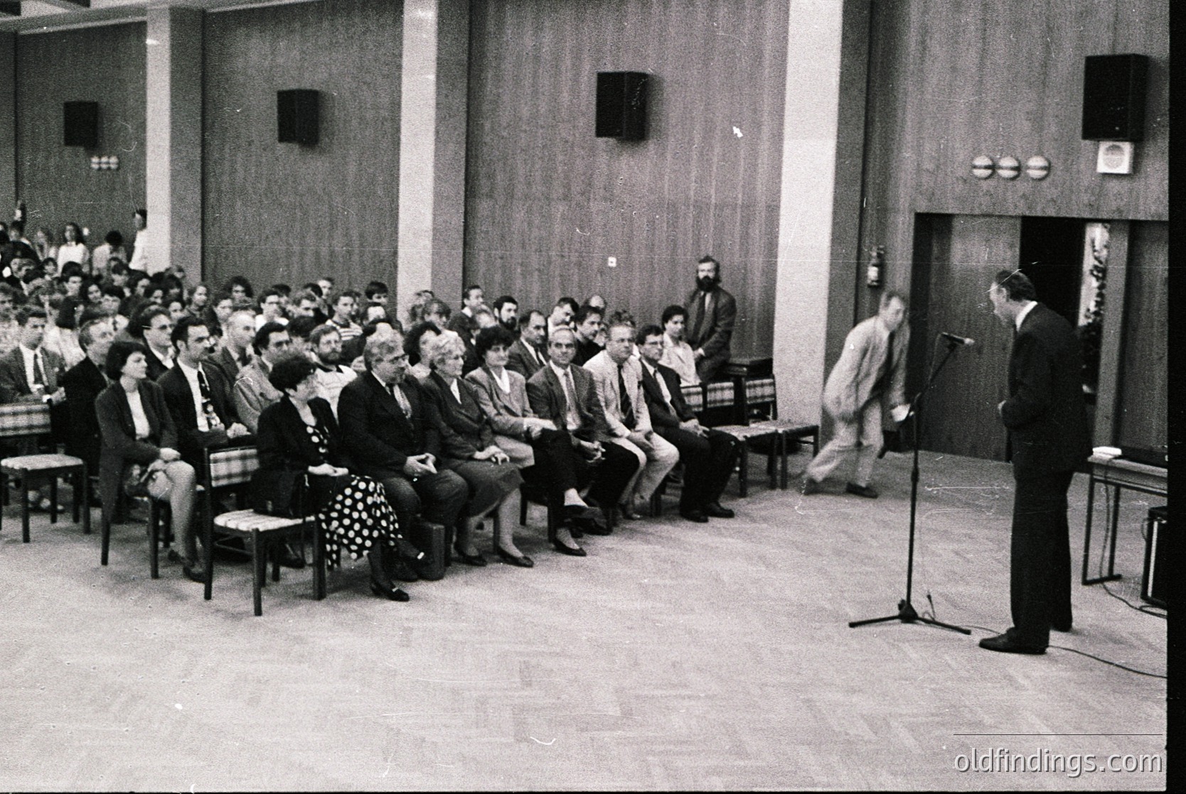 Mid-century lecture hall with tiered seating, featuring a speaker at a podium with microphone. Audience in formal attire, likely 1960s–1970s. Minimalist industrial design with exposed speakers and wooden paneling.