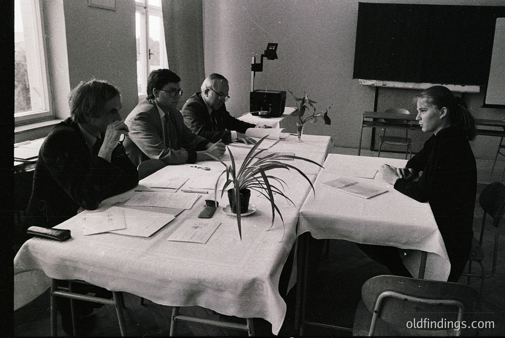 Four individuals seated around a rectangular table in a formal indoor setting, likely a conference or meeting room. The table is covered with white tablecloths, holding papers, a potted plant, and ink bottles. The men wear suits; the woman on the right wears a dark blazer. A projector screen and projector are visible in the background. Style and lighting suggest a mid-20th-century office environment. [Mid-20th-century office meeting with formal attire, projector screen, and papers on table ]