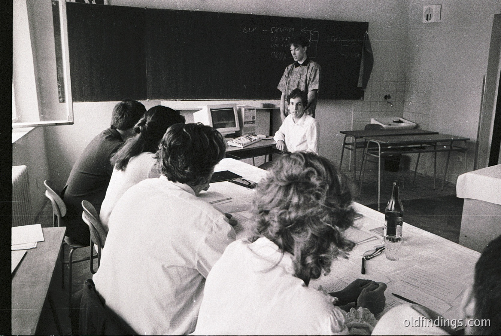 Classroom setting with students seated at desks, facing a teacher at a raised station. Blackboard with chalk writing, vintage typewriter, and basic office equipment visible. Likely late 20th century (1970s–1980s) educational environment.