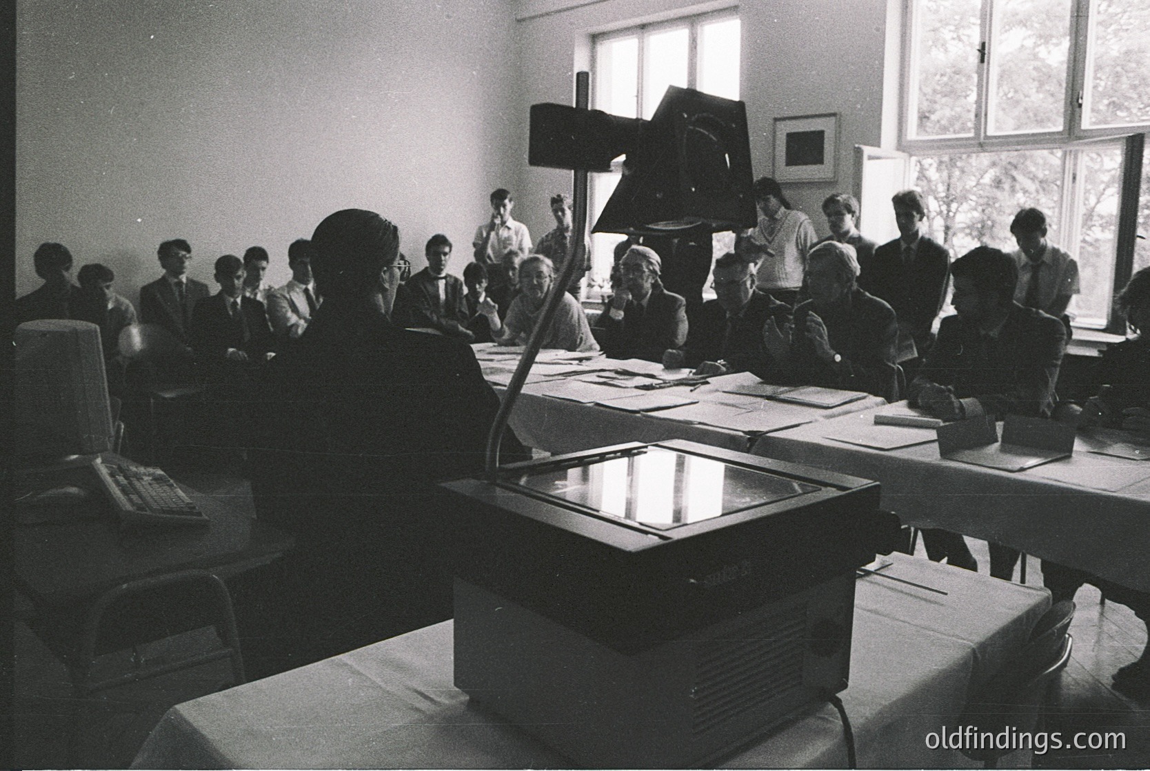Mid-20th century conference room with vintage projector and attendees in formal attire. Large screen reflects light, suggesting a presentation or lecture. Tables covered in white tablecloths, classic office chairs. Likely educational or corporate setting, 1950s–1970s.