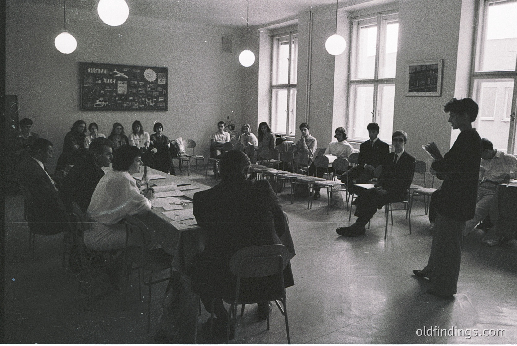 Mid-20th century classroom with U-shaped desks, students seated facing instructor. Blackboard displays solar system diagram. Hanging globe and industrial-style pendant lights. Formal attire suggests 1950s–1960s academic setting.