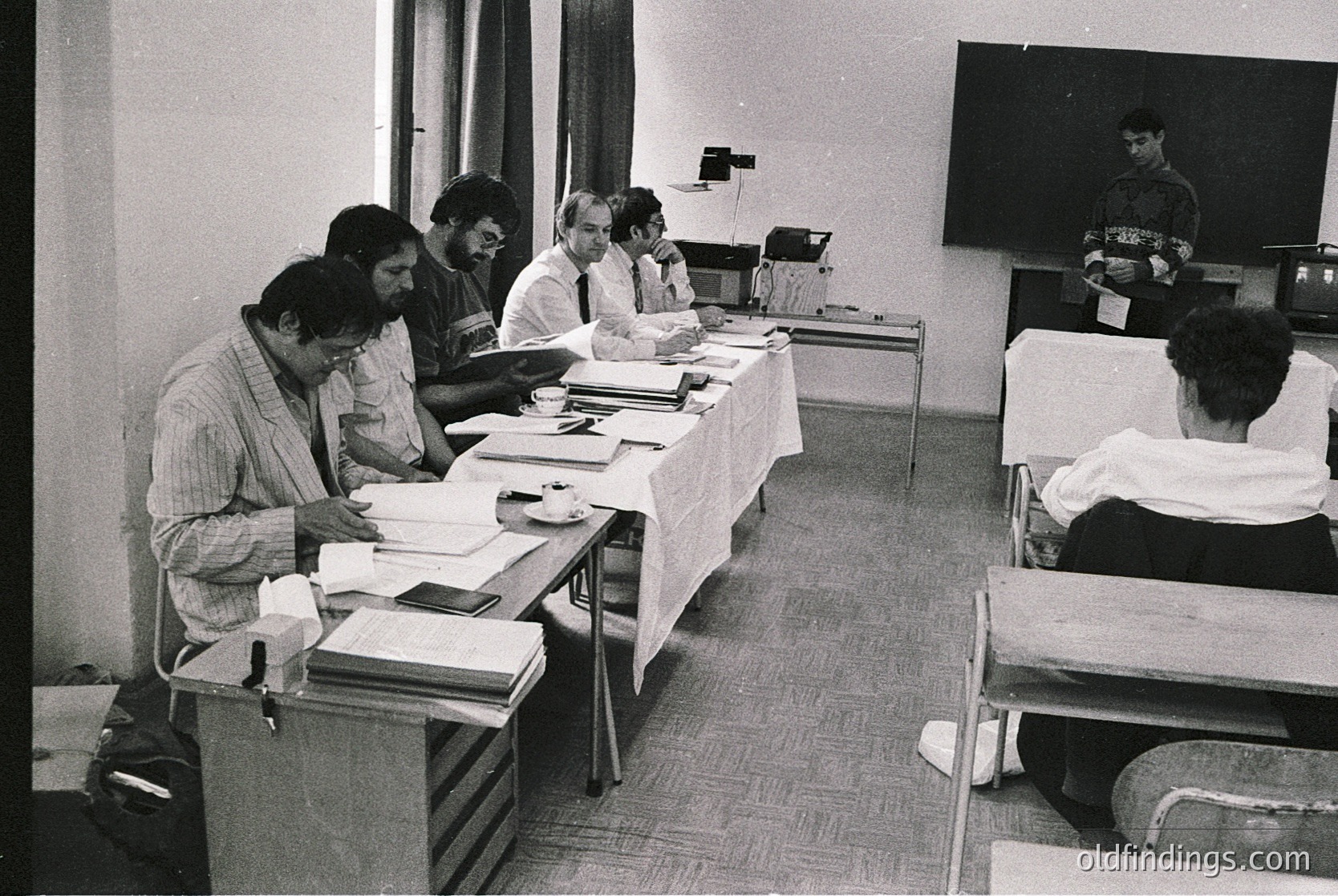 Black-and-white conference scene: six men seated at a rectangular table reviewing documents, surrounded by stacks of papers. Projector screen and projector on right wall. Casual office setting with a couch in background. Likely mid-20th century ( ) academic or corporate meeting.