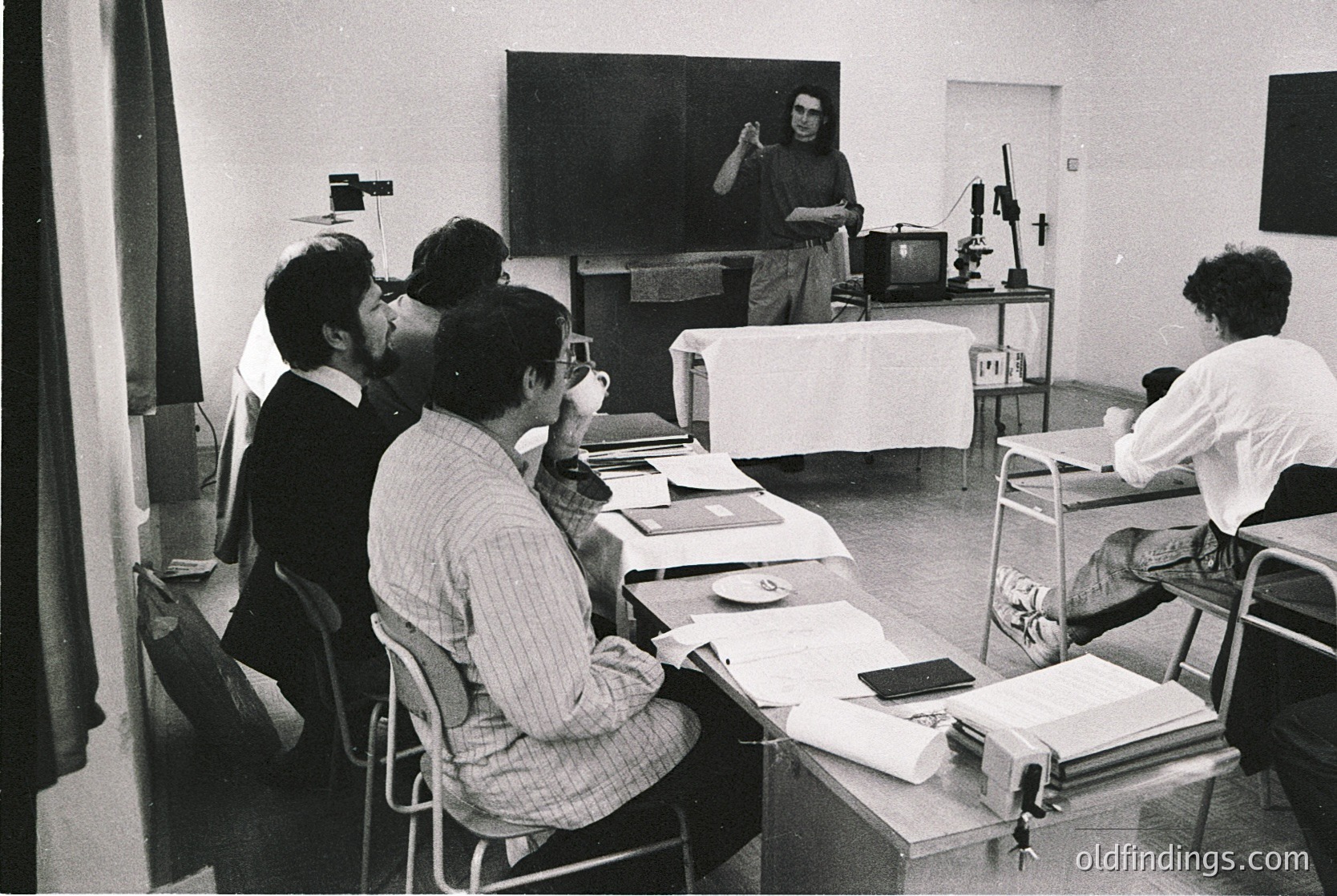 Black-and-white classroom scene featuring a lecturer gesturing at a chalkboard while students take notes, 1960s–1970s. Wooden desks, vintage chairs, and a projector setup suggest an academic or technical institution. Formal attire and minimalist decor reflect mid-century educational norms.