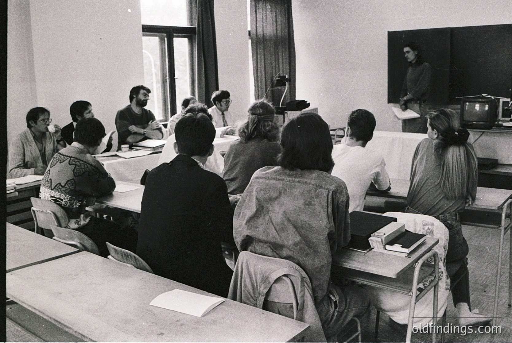 Black-and-white classroom scene with 12 seated students facing a lecturer at a podium. Wooden desks, vintage chairs, and a TV on a stand in the background suggest a 1970s-1980s educational setting. Formal attire and structured seating reflect institutional learning environments.