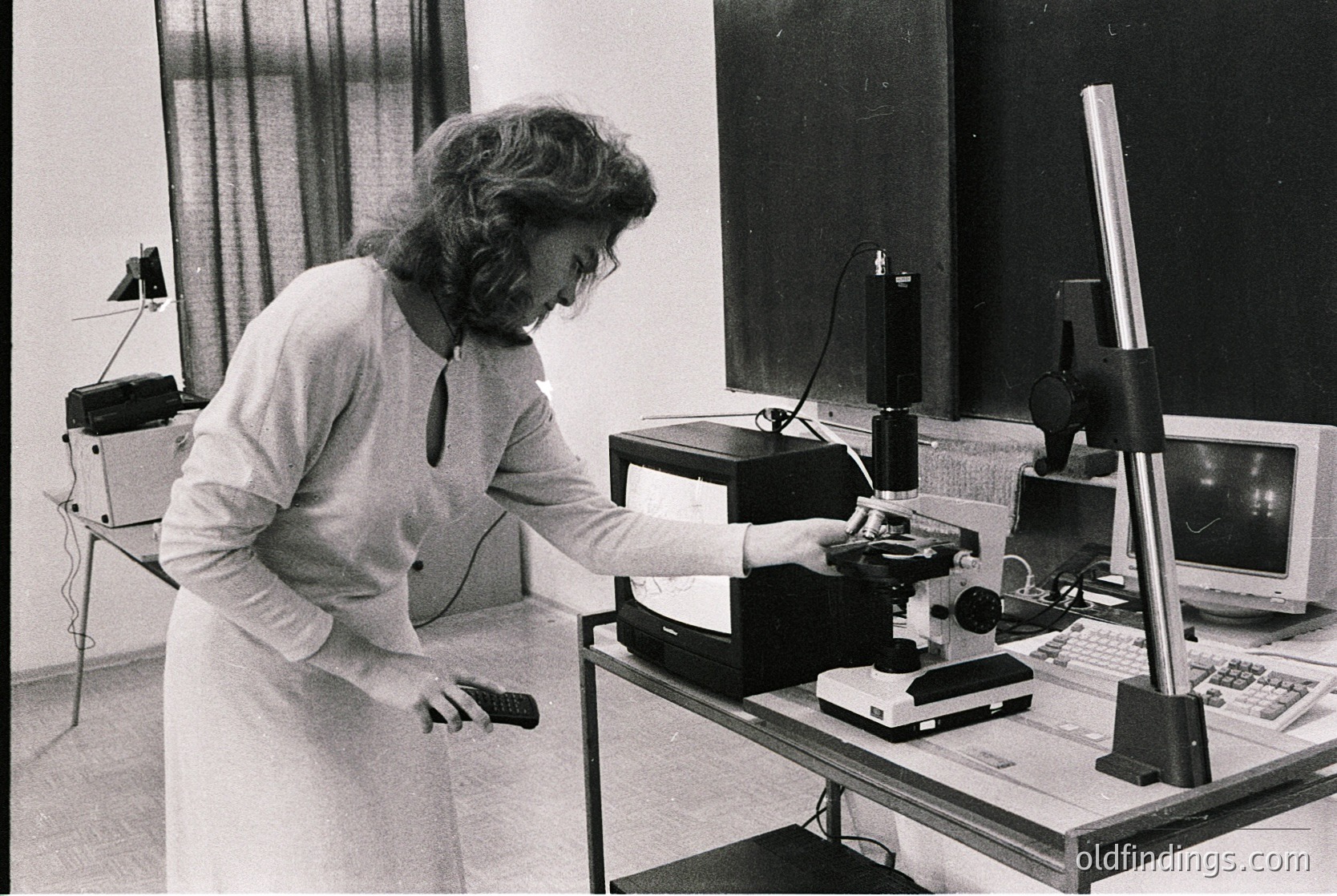 A professional woman in a lab coat operates vintage optical equipment, likely a microscope or slide projector, in a 1970s-era workspace. Surrounding tech includes a typewriter, reel-to-reel tape recorder, and early desktop computer. Industrial or academic setting.