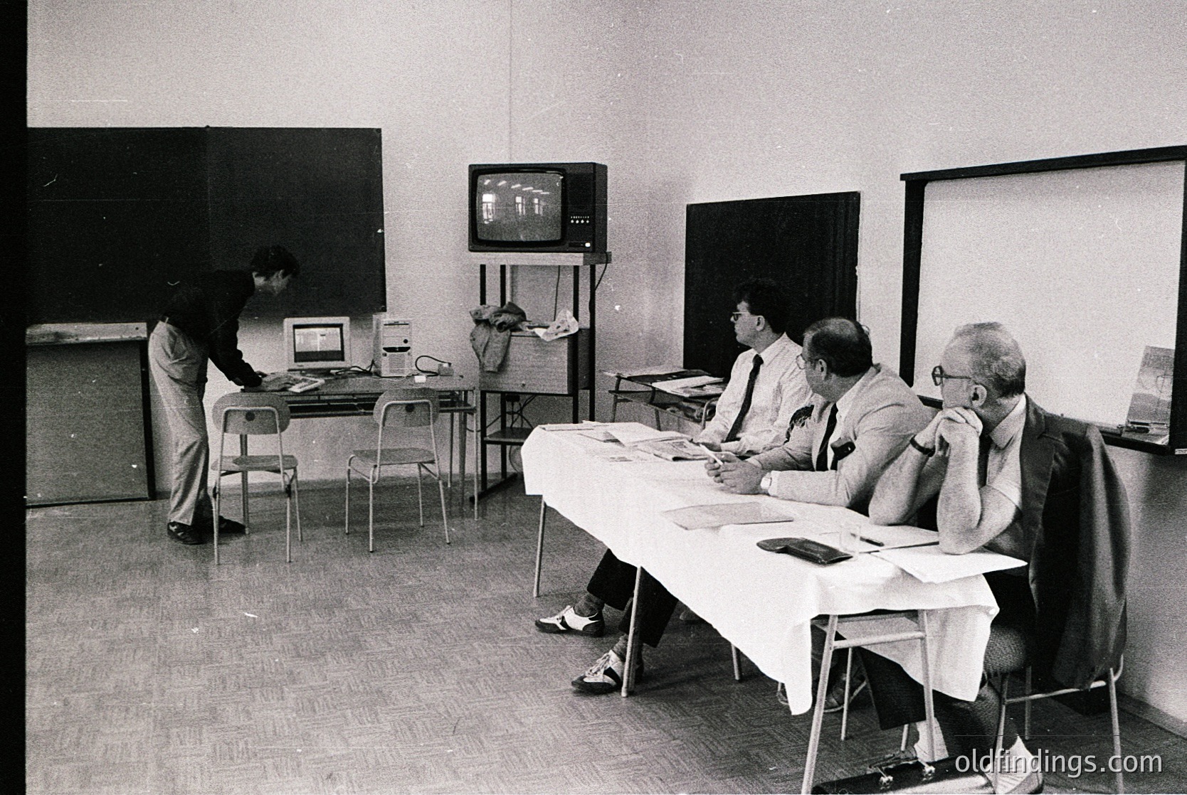 Vintage black-and-white office scene with four men in formal attire (1960s-70s). One man stands at a typewriter, others seated at a table with papers, under a TV displaying grainy footage. Minimalist industrial decor with chalkboard walls and metal chairs.