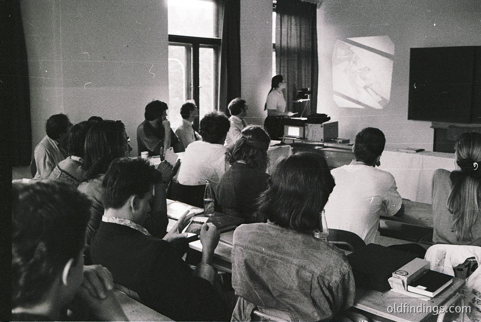 Black-and-white lecture hall scene from the 1960s–70s, featuring a speaker at a podium with projected slides. Audience faces forward, seated in tiered rows. Classic mid-century classroom design with wooden desks, natural light from windows, and a projector screen.