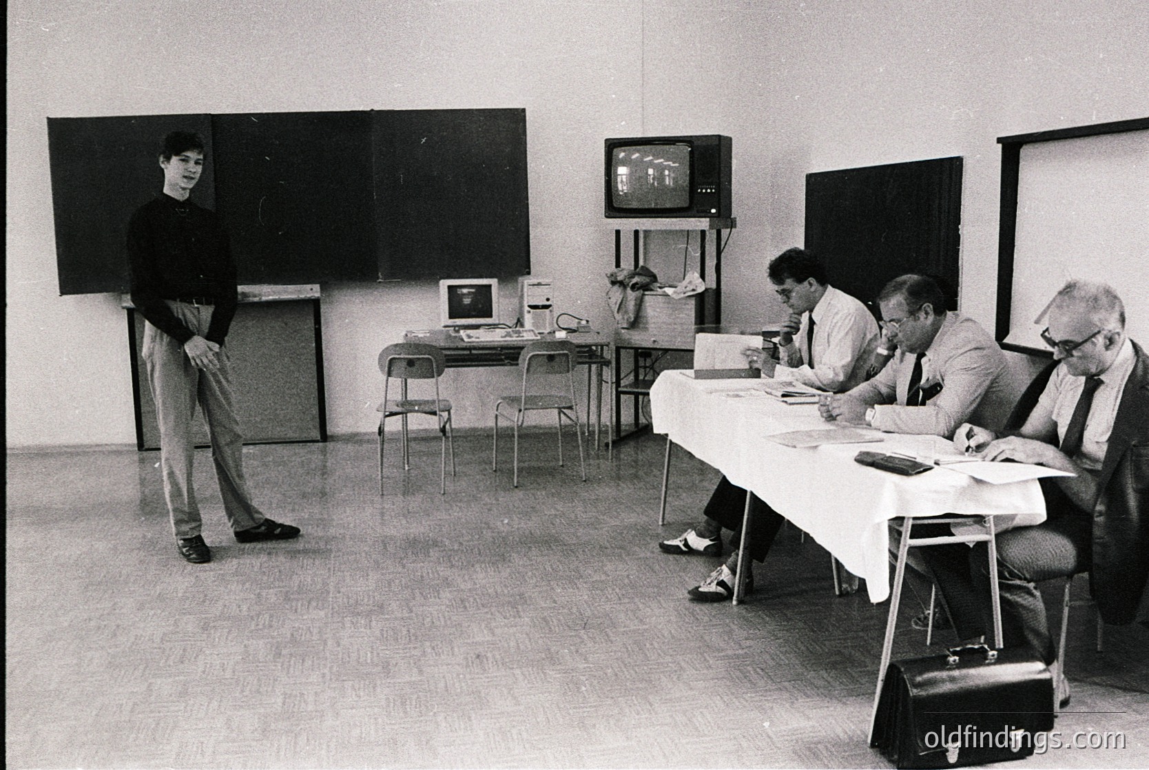 Black-and-white 1970s office scene: a man in a sweater stands beside a chalkboard, while four seated professionals review documents at a table. Early tech includes a CRT monitor and typewriter. Institutional setting likely educational or corporate.