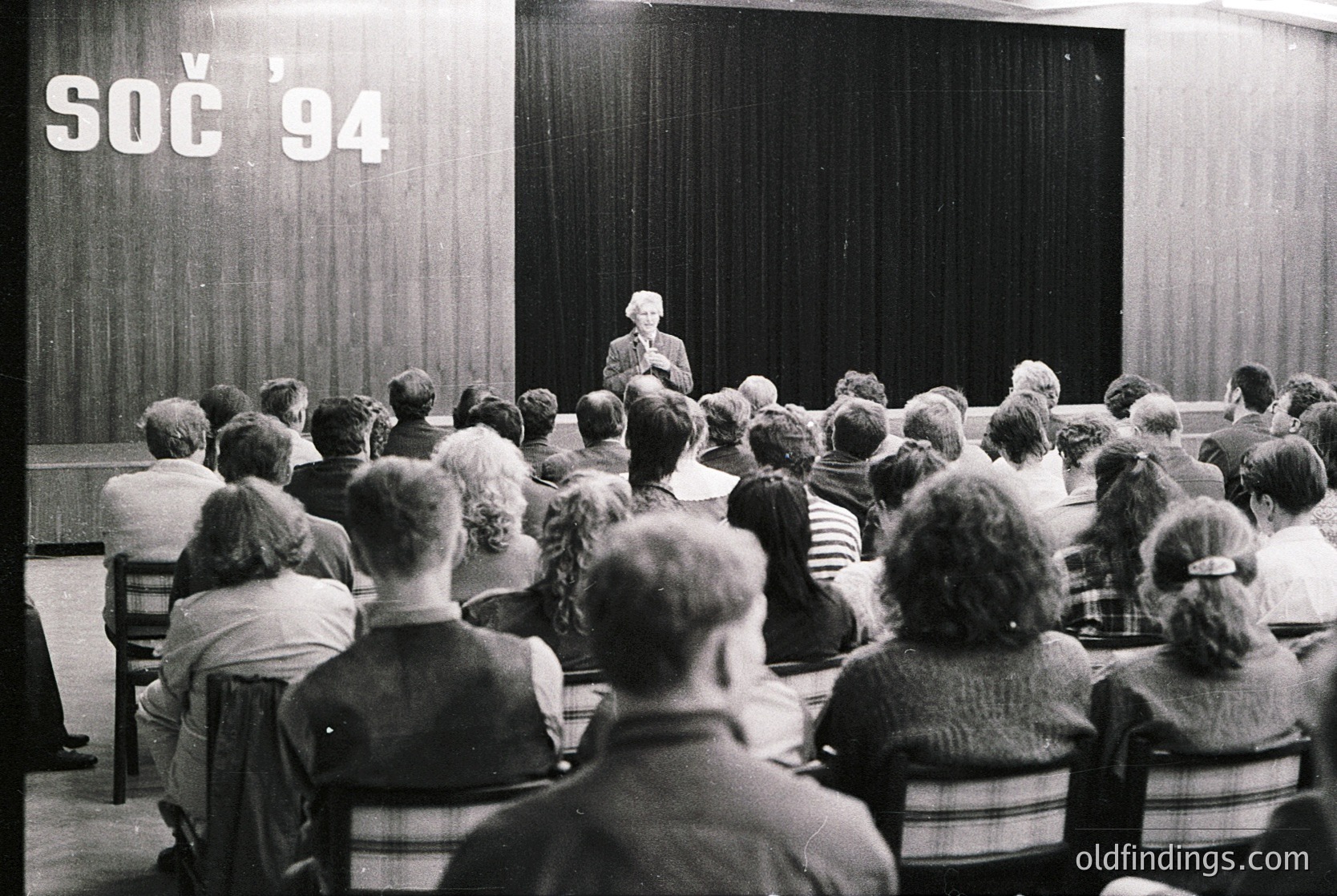 A black-and-white lecture hall scene from **Sochi 1994**, featuring a speaker addressing an attentive audience seated in tiered rows. The banner reads "СОЧ '94" (Sochi '94), indicating a conference or event. Formal attire and focused expressions suggest a professional or academic gathering.