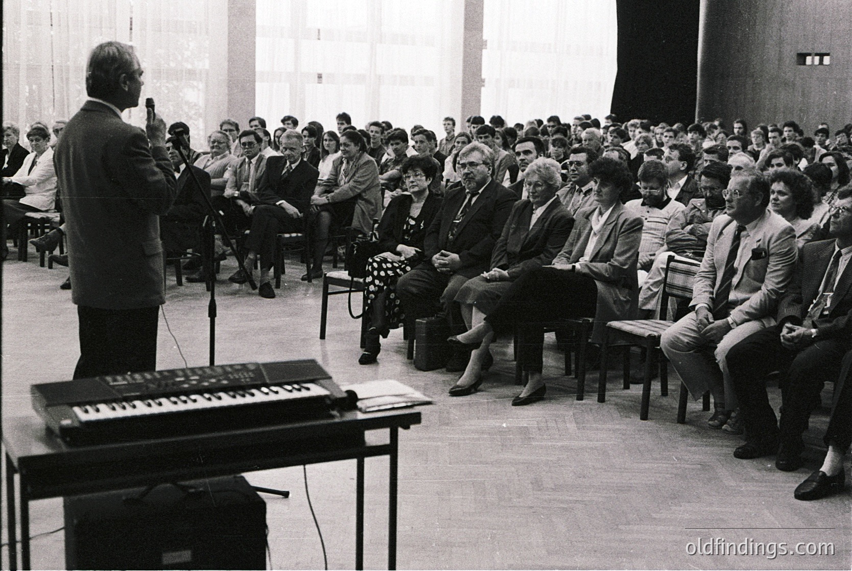 Black-and-white lecture hall scene featuring a speaker addressing a seated audience of ~80 individuals in formal attire (1960s–1970s). Stage setup includes a vintage keyboard and microphone stand. Spacious hall with tiered seating and minimal decor.