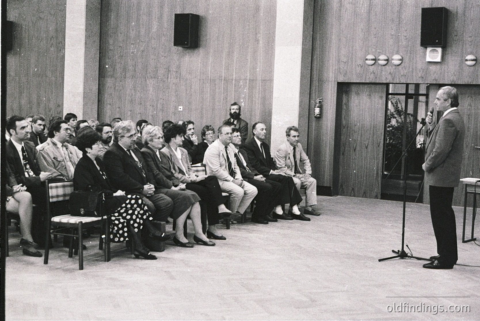 A formal indoor gathering in a mid-century conference hall, featuring a speaker addressing an attentive audience seated in wooden chairs. The setting includes wood-paneled walls, a central speaker’s podium, and two large speakers mounted on the walls. Attendees wear 1960s–70s formal attire, suggesting a professional or governmental event. The lighting and composition evoke a structured, institutional atmosphere.