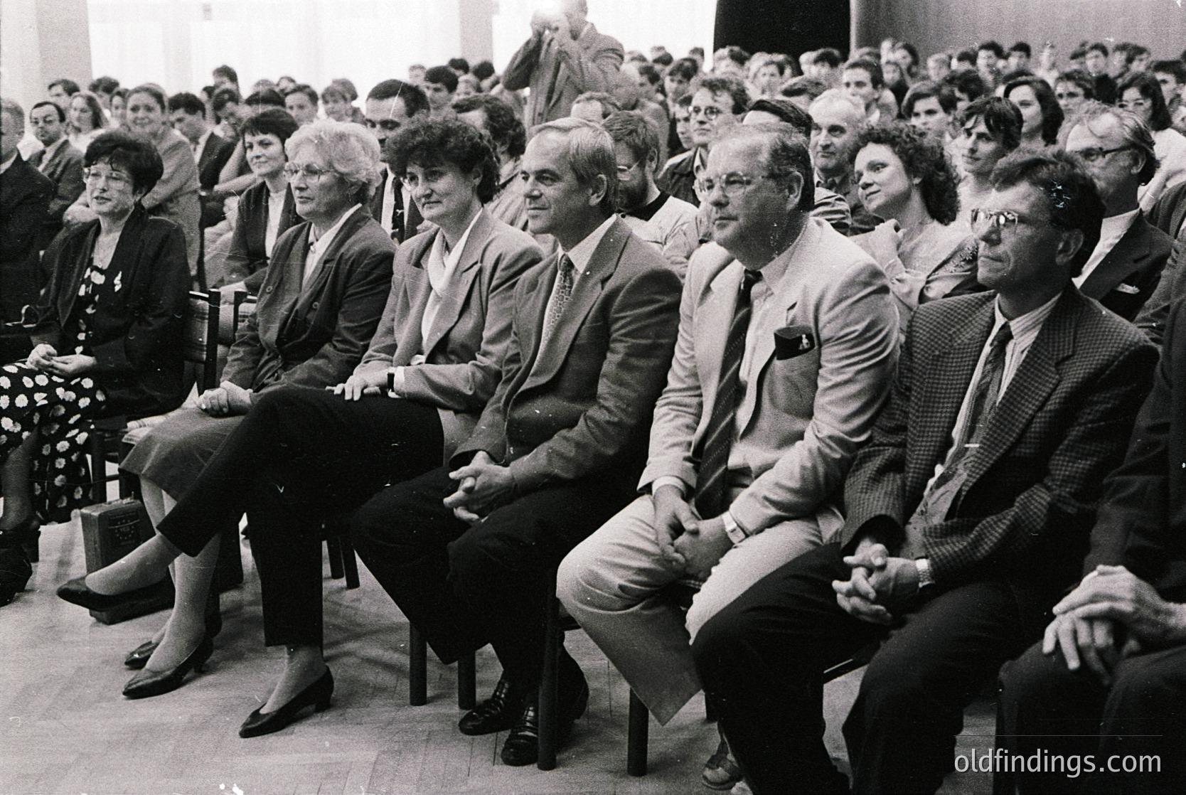 1970s indoor gathering of formal attire attendees in a packed auditorium. Wooden chairs, tiered seating, and a stage backdrop suggest a conference or official event. Attire includes blazers, ties, and dresses—indicating professional/corporate or governmental context.