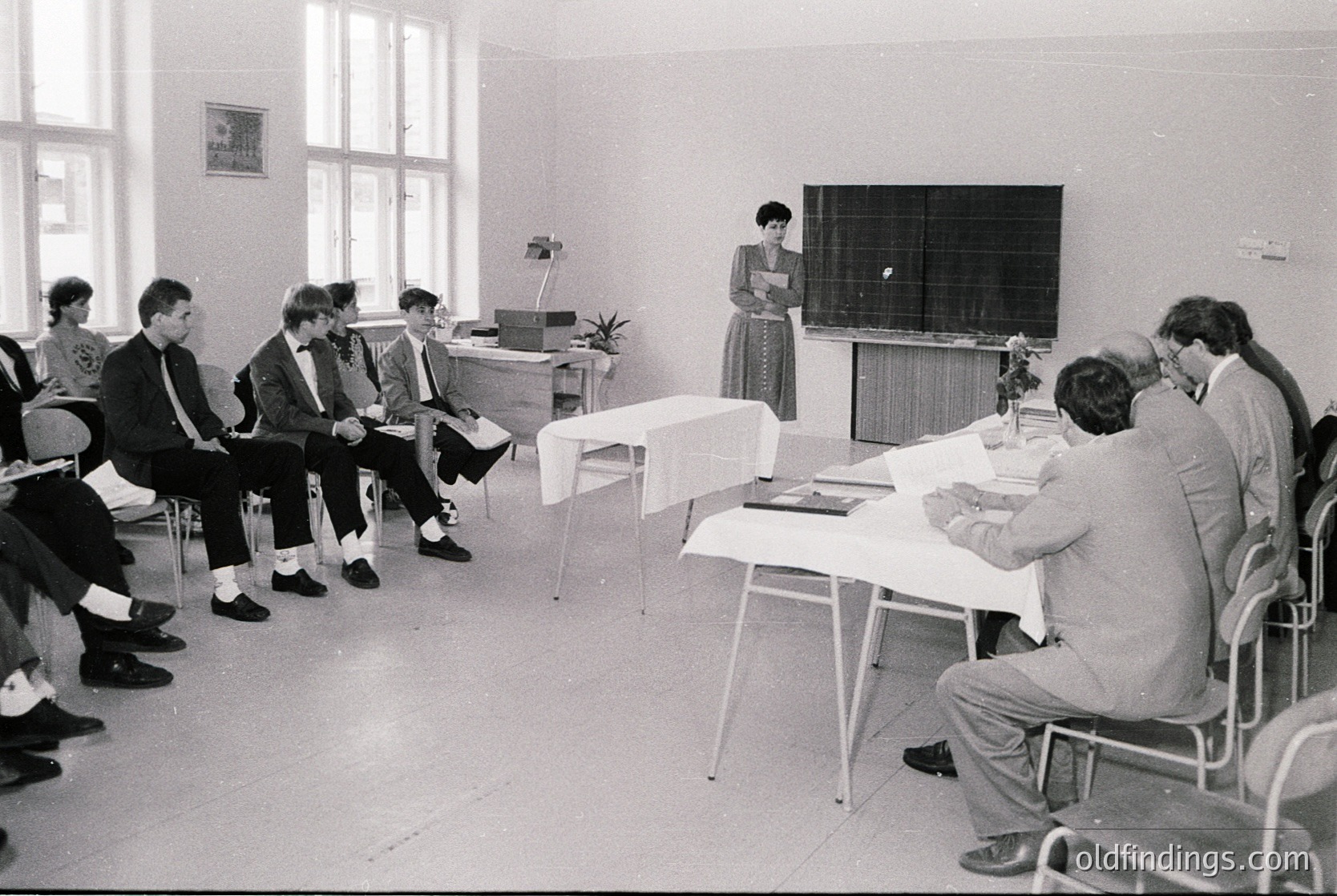Mid-20th century classroom seminar with 12 seated attendees in formal attire, facing a standing presenter at a lectern. White desks, metal chairs, and minimalist decor suggest institutional education. Likely Eastern Bloc-era institutional setting (, ).