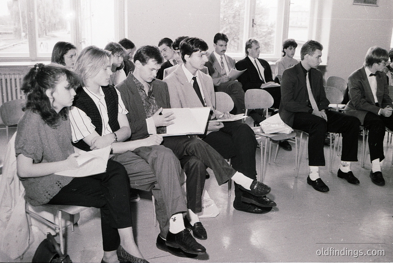 Classroom setting featuring 1970s-era students in formal attire—mixed-gender group seated on plastic chairs, reading papers or notebooks. Institutional lighting and basic furnishings suggest an educational institution. Likely Eastern Bloc or Soviet-era due to attire and setting.