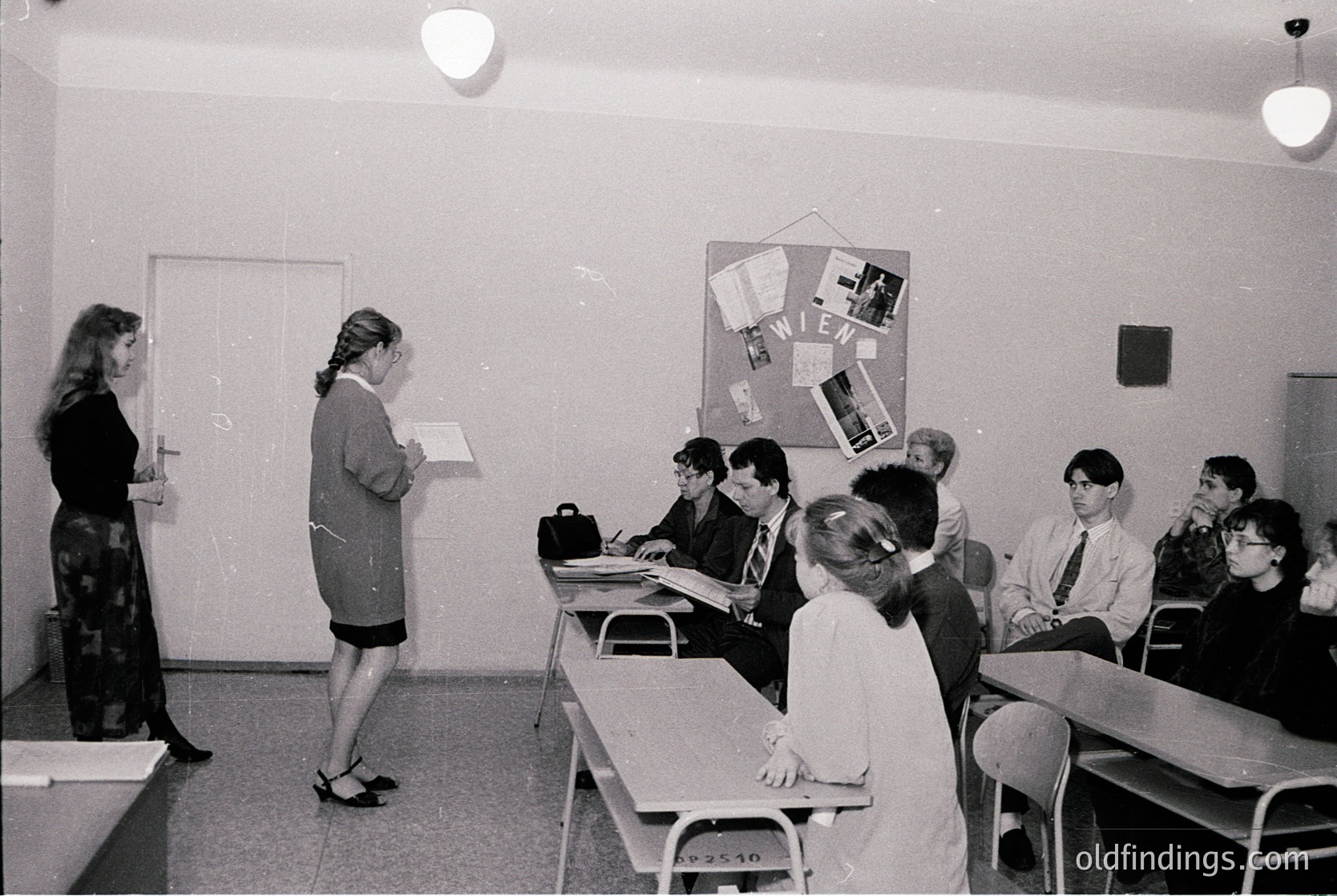 Spacious classroom with 1970s/80s design—teacher in skirt suit directs students seated at desks. Bulletin board displays "VIEW" with pinned papers. Fluorescent lighting and metal chairs. Likely Eastern Bloc-era educational setting.