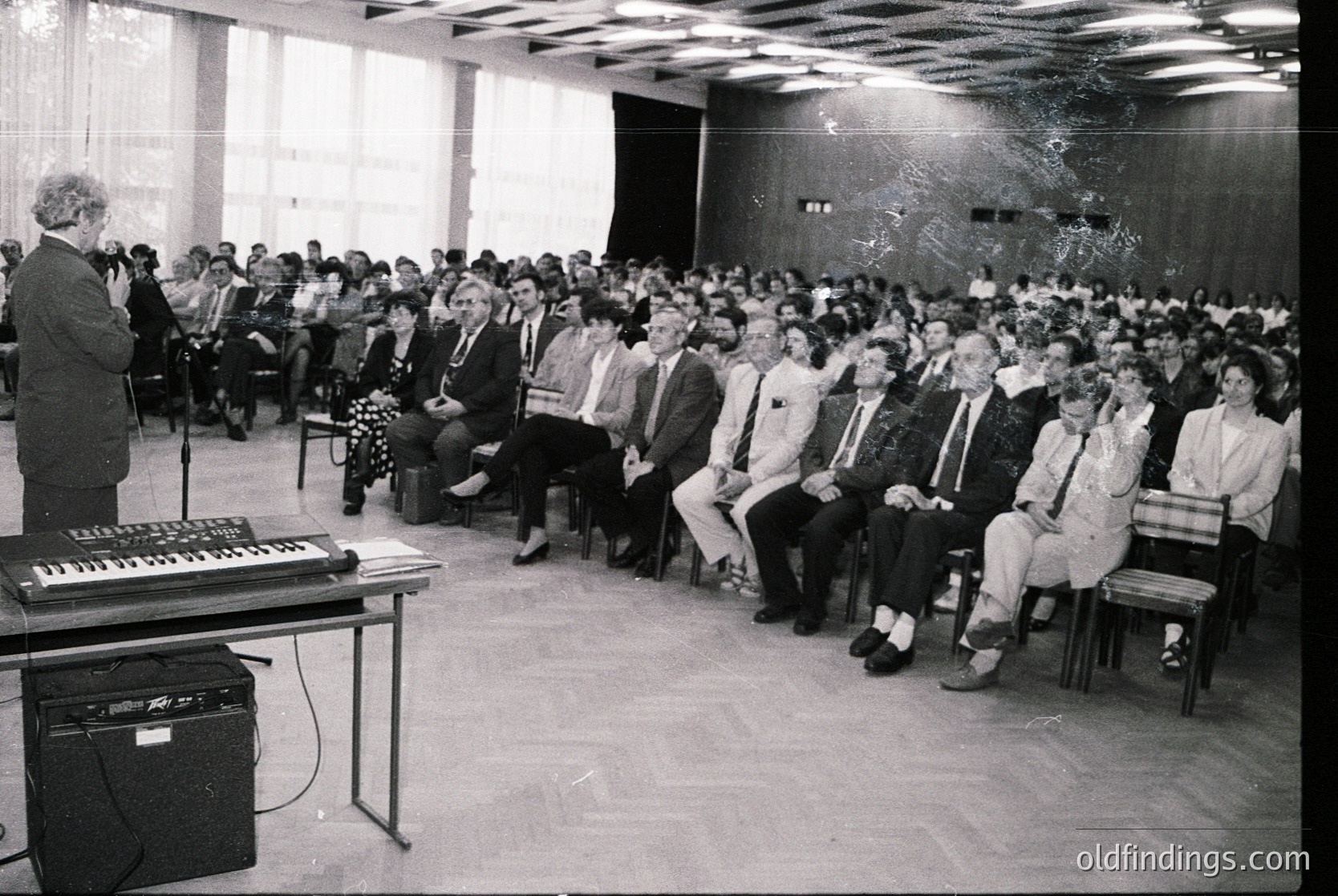 A black-and-white lecture hall scene from the 1970s–1980s, featuring a speaker at a podium with a vintage keyboard and amplifier. Audience of ~100 seated in tiered rows, dressed in formal attire (blazers, ties, dresses). Minimalist concrete architecture with exposed ceiling pipes. Likely an academic or professional event.