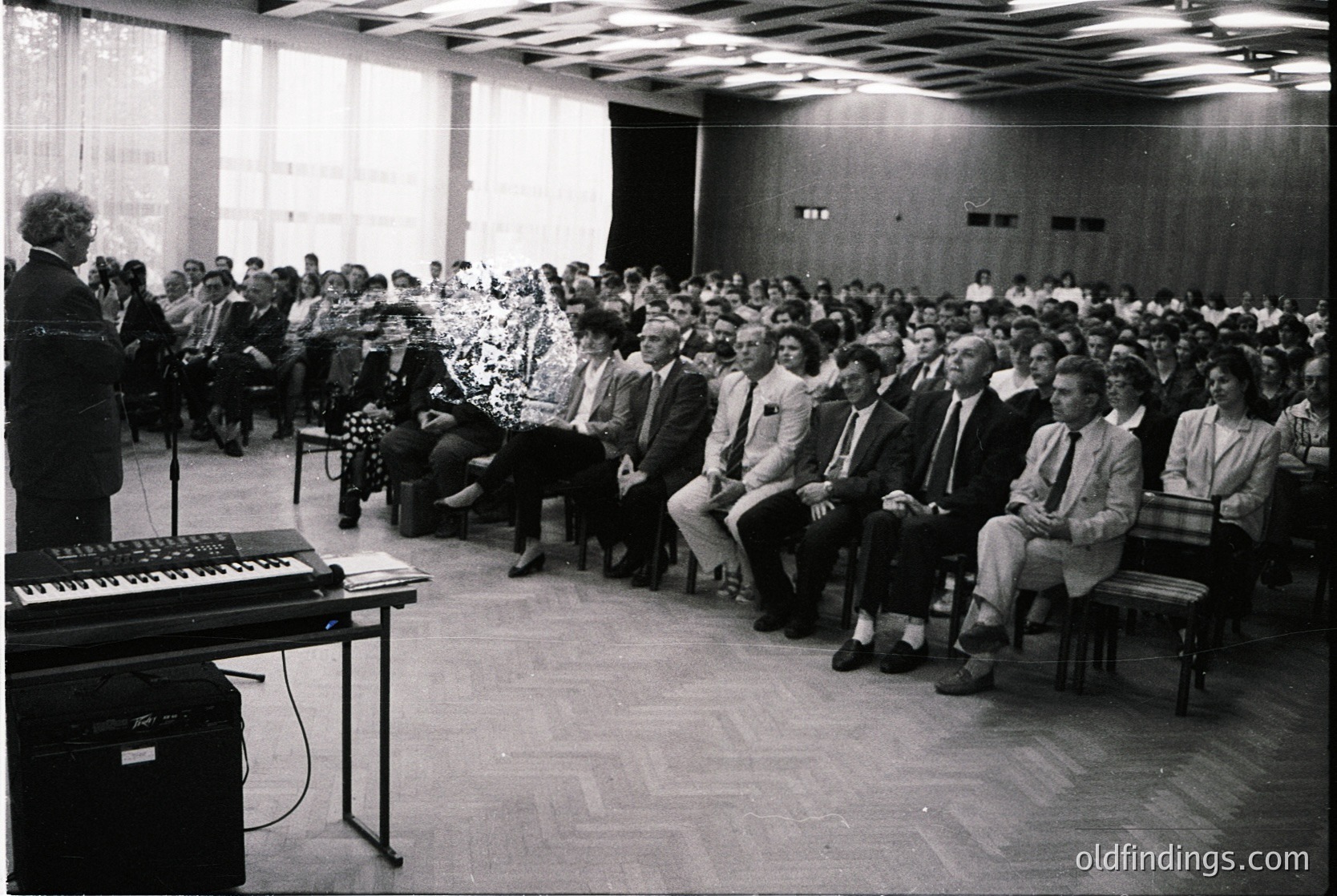 Mid-century lecture hall with tiered seating, featuring a speaker at a podium with a keyboard instrument (likely a Wurlitzer). Audience of ~100+ dressed in formal attire (men in suits, women in dresses), suggesting a professional or academic event. Decor includes floral centerpieces on tables and a curtained window. Likely Eastern Bloc-era institutional setting ( ).