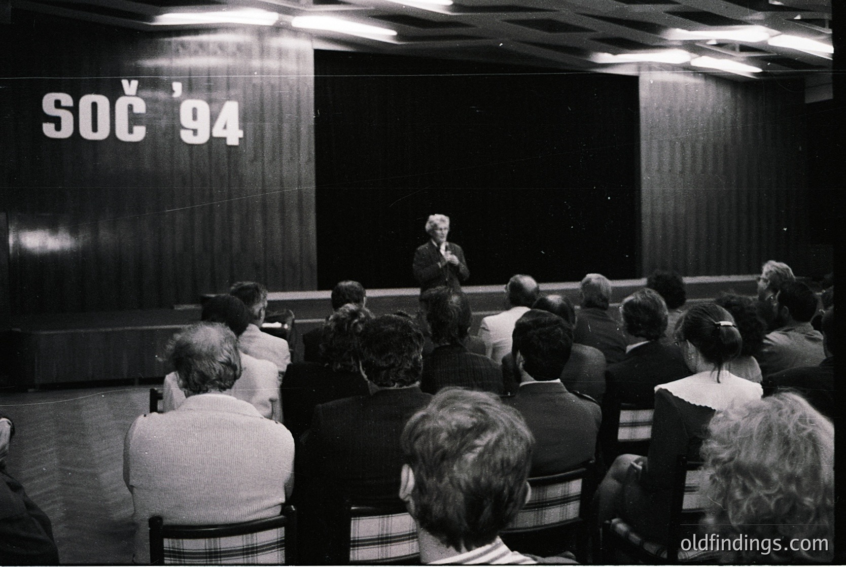 Black-and-white lecture hall shot at the 1994 Summer Olympics (SOC) in Lillehammer, Norway. Speaker addresses seated audience in tiered seating, with "SOC '94" banner prominently displayed. Formal attire suggests official event or press conference.