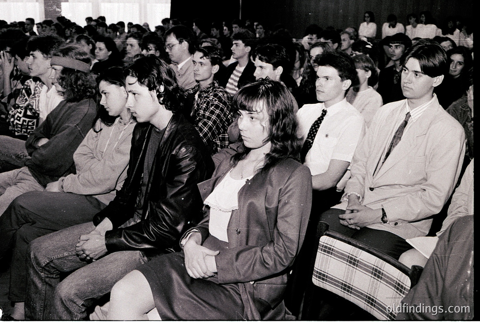 Black-and-white shot of a packed indoor lecture hall, likely from the **1970s-1980s**. Audience of young adults in formal attire—men in dress shirts, ties, and plaid jackets; women in blazers and blouses—sitting attentively. Crowded seating suggests an academic or professional event. Formal setting hints at structured, institutional gatherings.