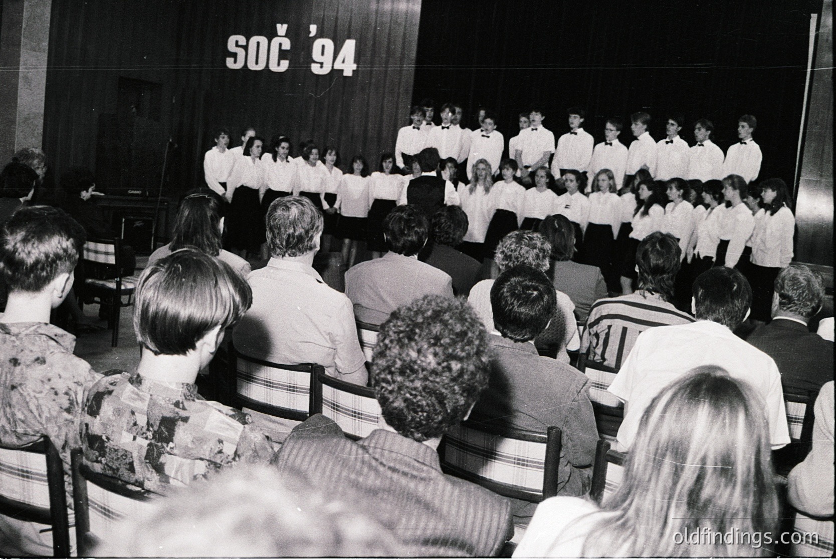 Large indoor choir performance at the 1994 Summer Universiade (SOC '94) in Fukuoka, Japan. Uniformed singers on stage, audience seated facing them. Black-and-white archival photo captures 1990s cultural event energy.
