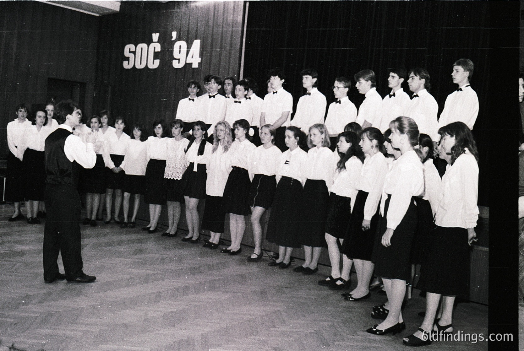 School choir rehearsal, 1994, likely Eastern Europe. Uniformed students in white blouses and dark skirts/shorts stand in rows, led by a conductor in dark suit. Indoor auditorium with wooden paneling and "SOC '94" banner.