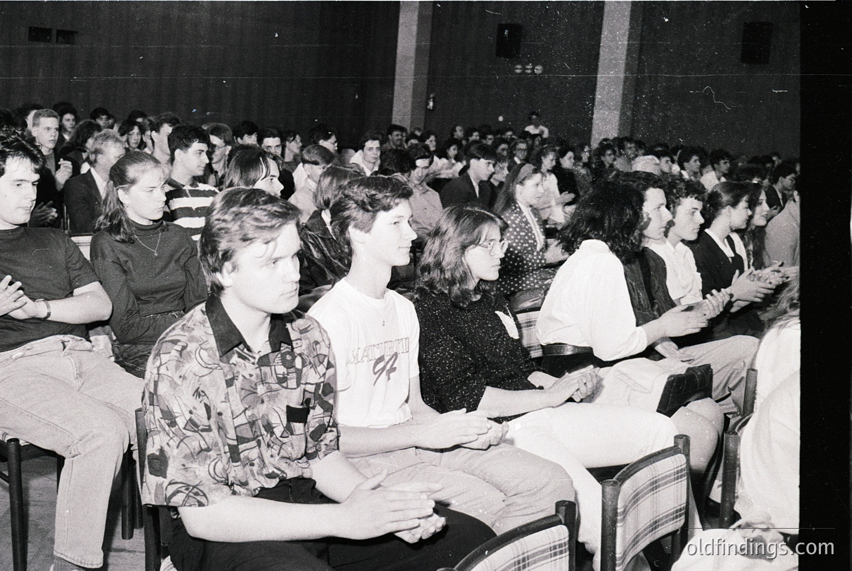 Black-and-white indoor lecture hall filled with seated students, likely 1970s–1980s. Wooden benches and tiered seating create depth. Foreground features a man in a patterned shirt and a woman in a graphic tee. Crowded atmosphere suggests academic or public event.