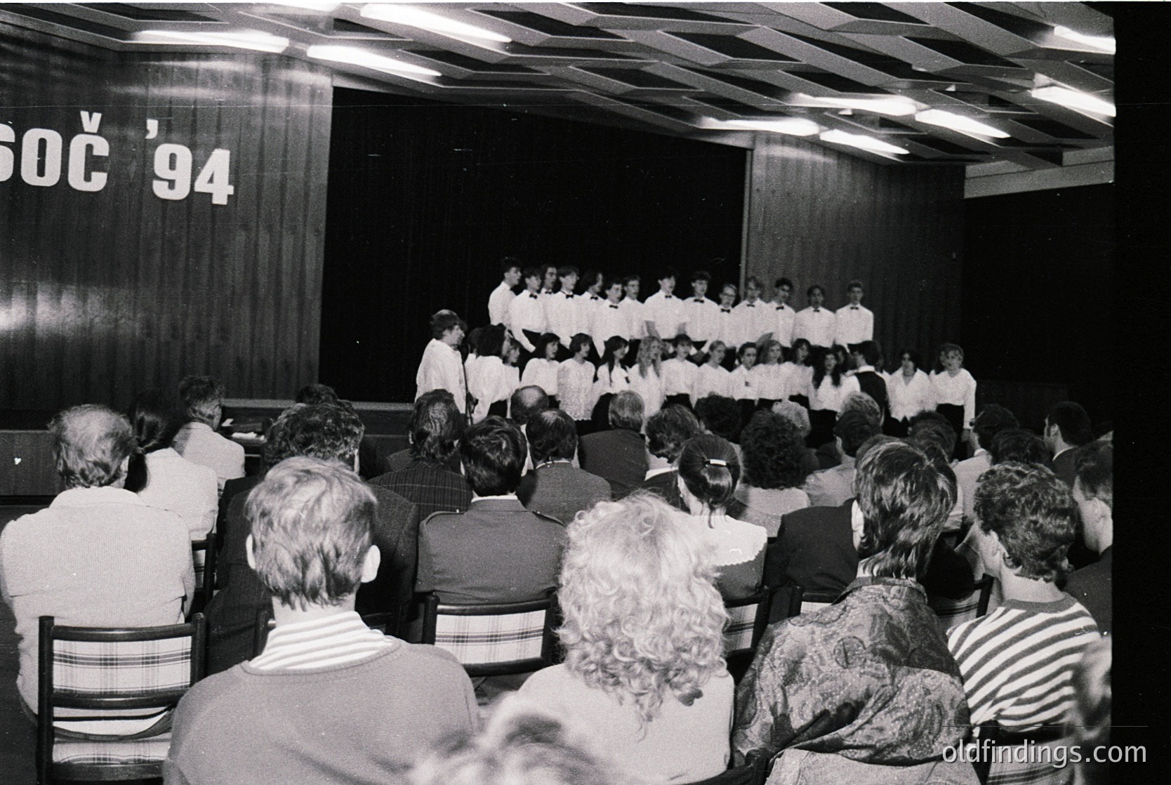 A black-and-white concert hall scene from **SOC '94** (likely the 1994 Song Contest in Croatia). A large choir in matching white shirts performs on stage, while an attentive audience faces them. The venue’s industrial-style ceiling and dated seating reflect 1990s Eastern European cultural events.