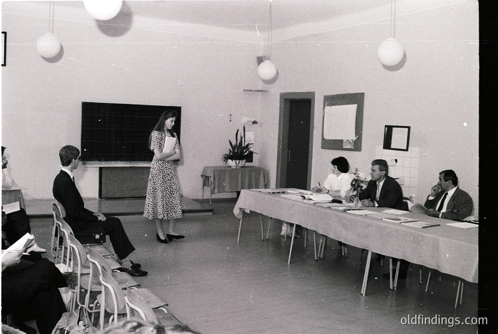 Mid-20th century classroom/conference setting with vintage 1960s-70s decor. Woman in floral dress addresses seated men at a long table; chalkboard and projector screen in background. Suspended globe lamps and potted plant add detail. Formal attire suggests institutional or professional event.