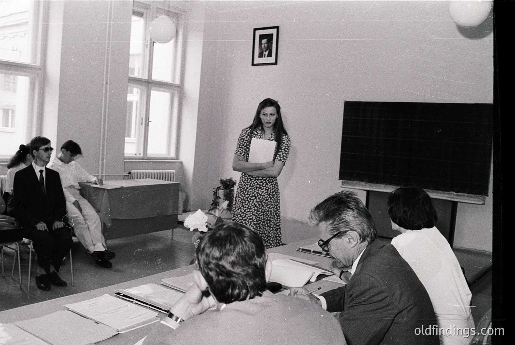 Mid-20th century classroom setting with formal attire and vintage teaching aids. A woman in a patterned dress stands at a chalkboard, addressing seated students and faculty. Desks arranged in rows, framed portraits on walls, and a projector screen hint at institutional education. Likely Eastern Bloc or Soviet-era academic environment.