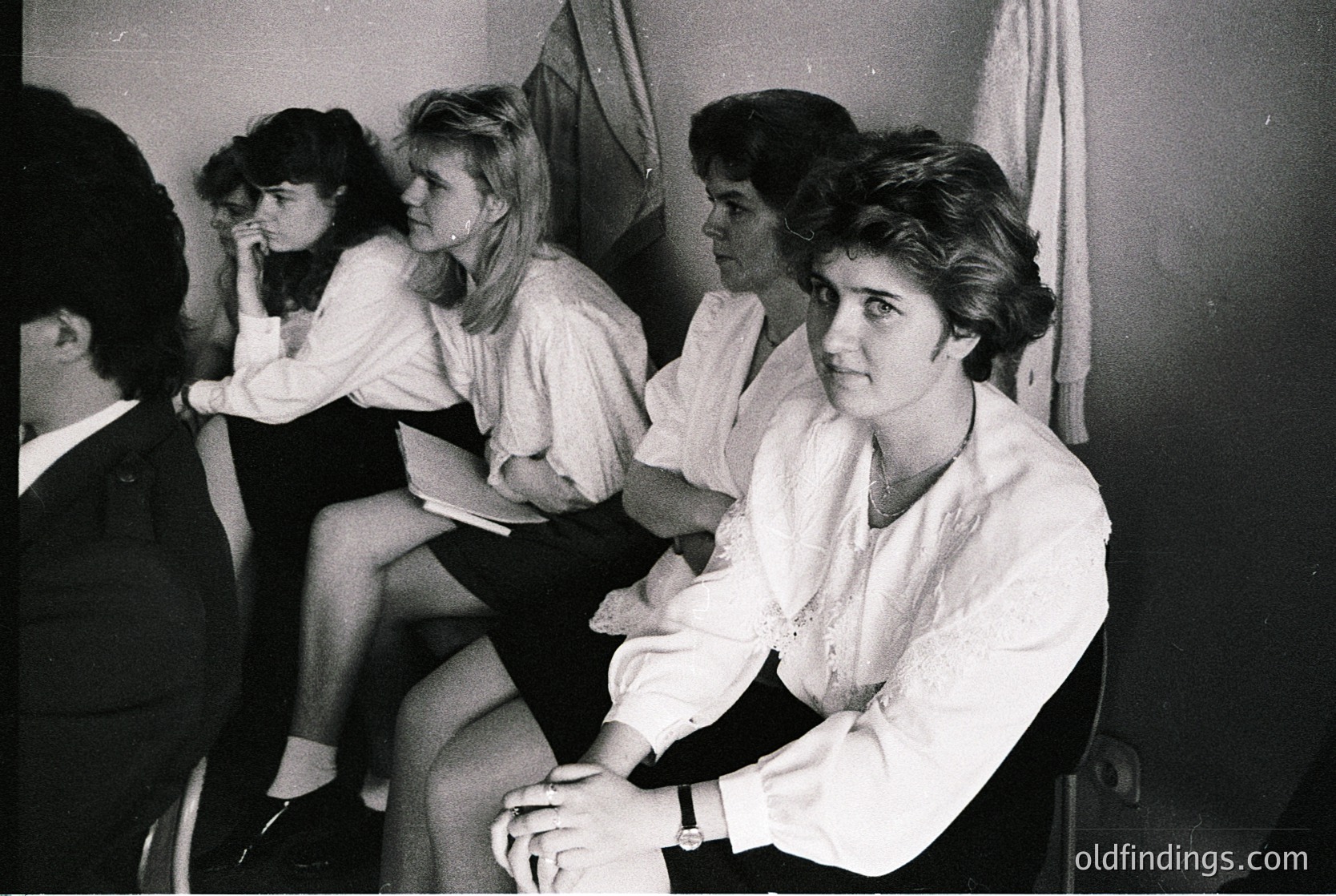 Group of four women in 1960s-era formal attire—blouses, jackets, and short hair—sitting in a dimly lit indoor setting, possibly a lounge or waiting area. One woman leans forward, others pose casually. Watch on wrist suggests mid-century fashion.