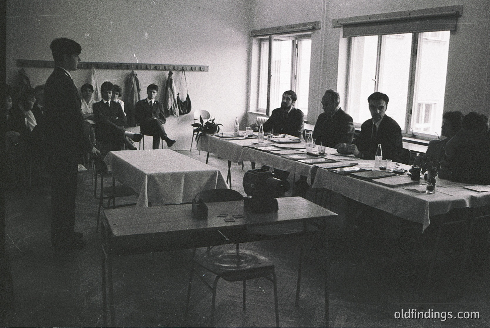 Classroom setting with vintage typewriters, likely mid-20th century. Six seated students and a teacher at desks with white tablecloths, writing materials, and vintage typewriters. High windows allow natural light. Formal attire suggests structured education environment.