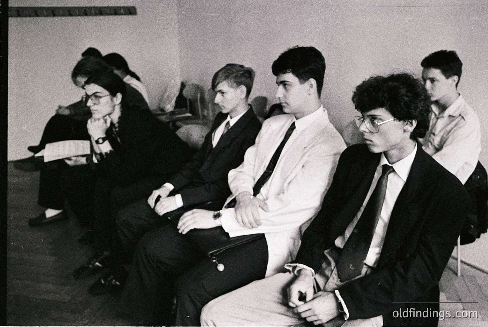 Black-and-white group portrait of five seated individuals in formal attire—men in suits, ties, and dress shirts; one woman in glasses and a headscarf. Plain indoor setting with wooden chairs and neutral walls, suggesting a 1960s–1970s institutional or academic environment.