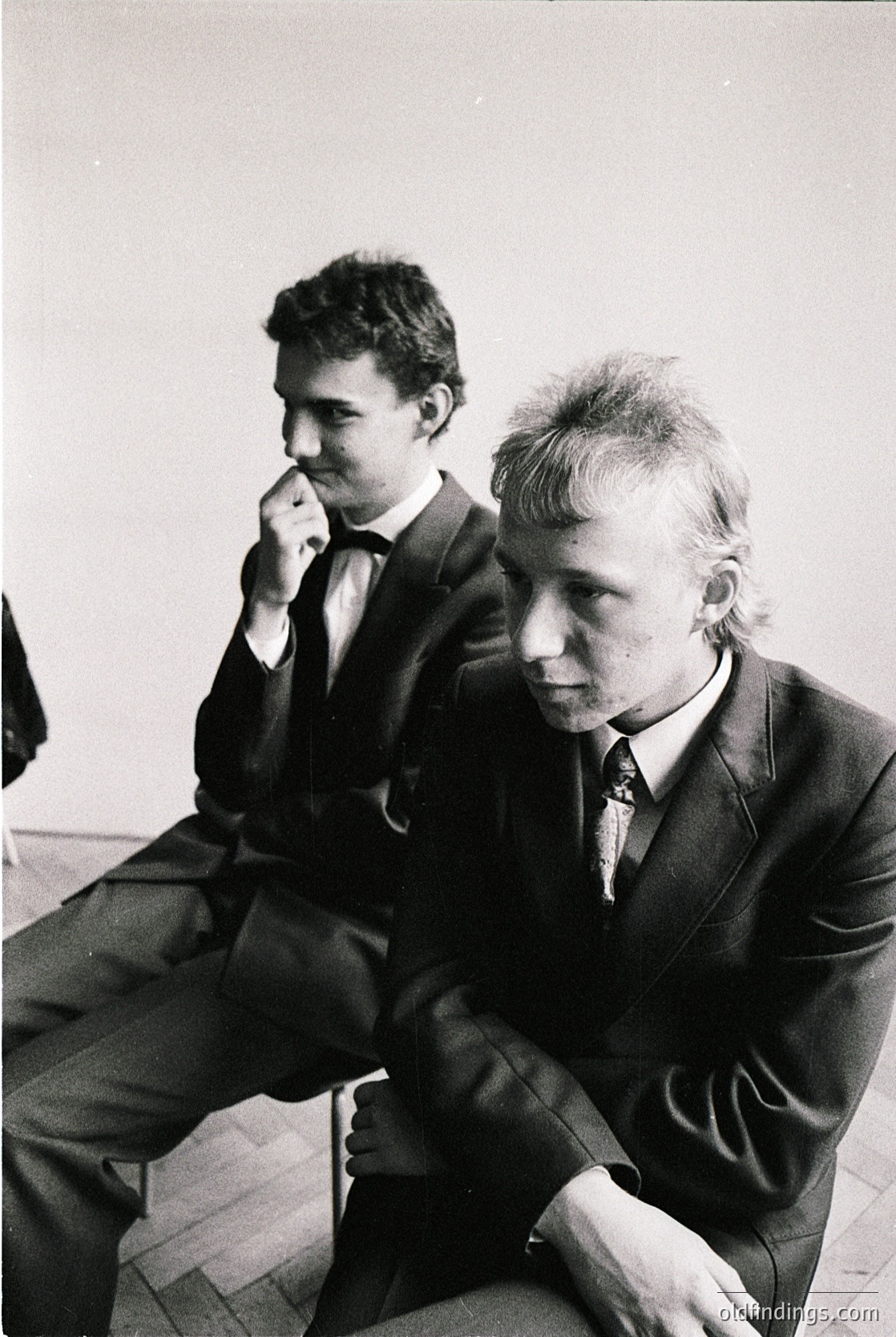 Two men in formal attire sit on stools in an indoor setting, likely a 1970s European office or meeting space. The man on the left wears a vest and tie, while the other sports a suit with a patterned tie. Both exhibit thoughtful expressions, suggesting a serious discussion or waiting period.