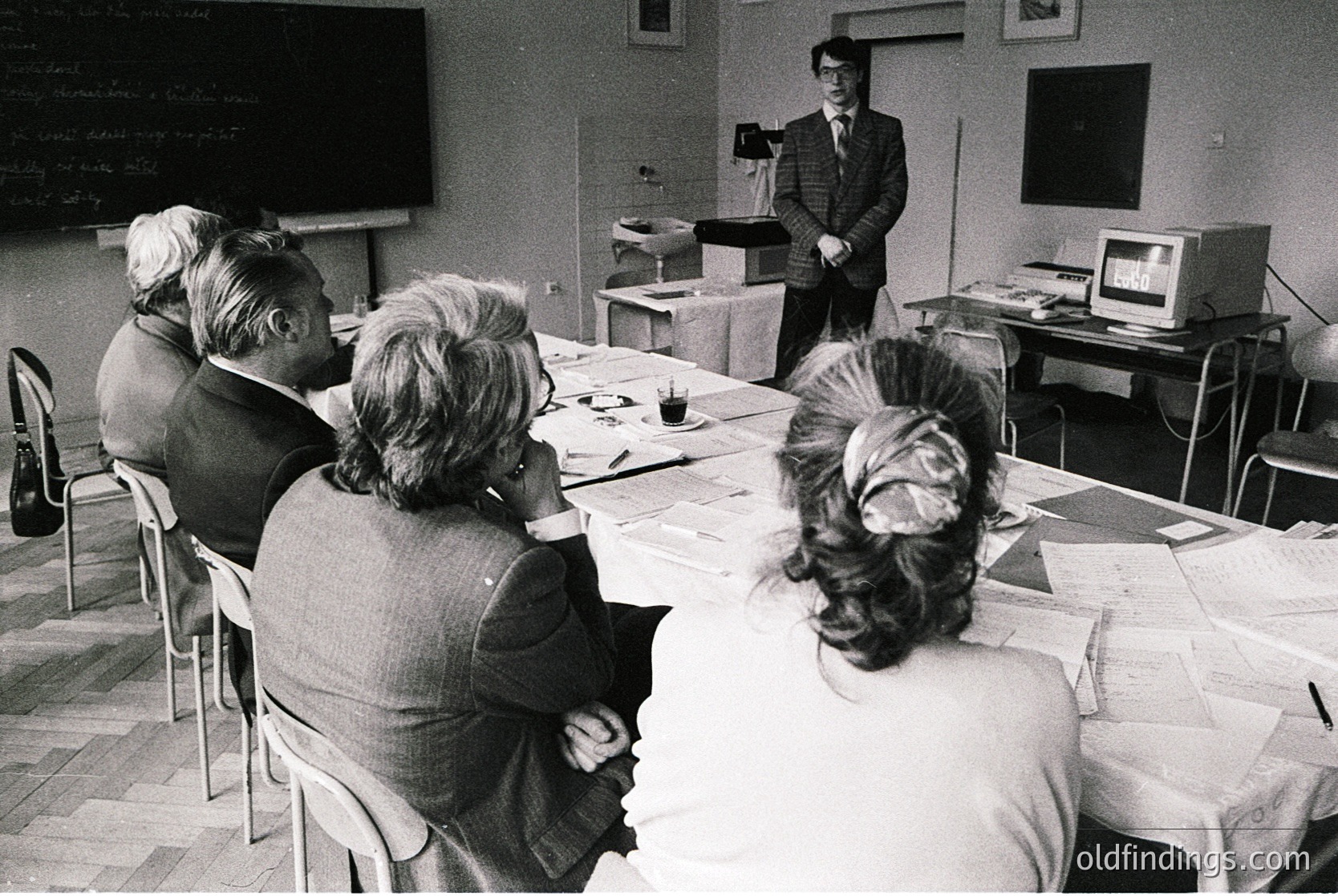 A 1970s-era classroom lecture featuring a standing instructor in formal attire addressing seated students. Tables covered in papers, a chalkboard with diagrams, and a vintage computer terminal in the background. Formal, institutional setting likely in a Western European or North American university.