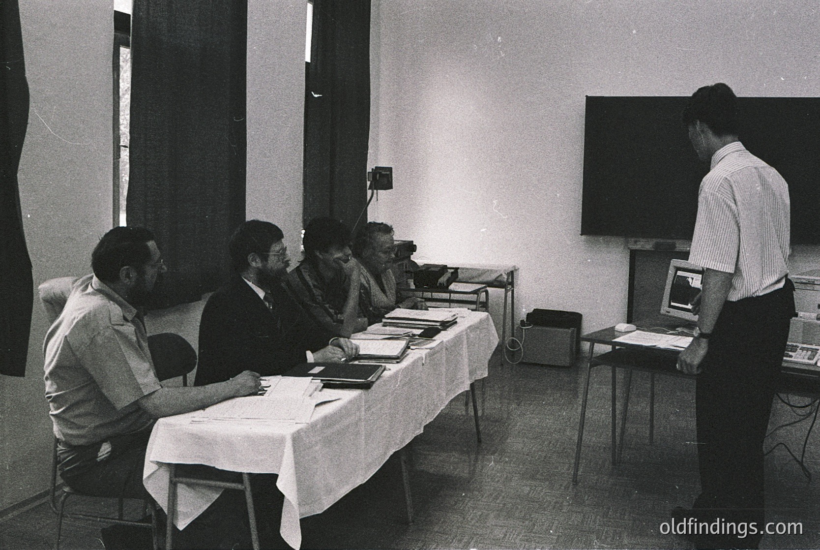 Black-and-white office scene from the 1970s–1980s, featuring five men in formal attire seated around a table with documents. One man stands at a projector setup, likely presenting slides. Minimalist decor with white tablecloths, basic lighting, and a projector screen. Ideal for historical research on corporate meetings or educational presentations.