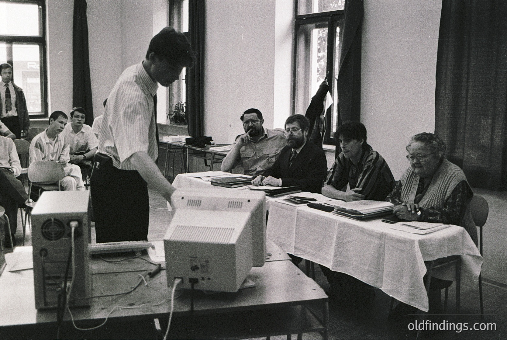 Early 1980s classroom featuring a vintage **Commodore PET 2001** computer. Group of adults—likely educators and students—engaged in hands-on computing. Industrial-era classroom setup with desks, white tablecloths, and a chalkboard in background. Reflects early adoption of personal computing in education.