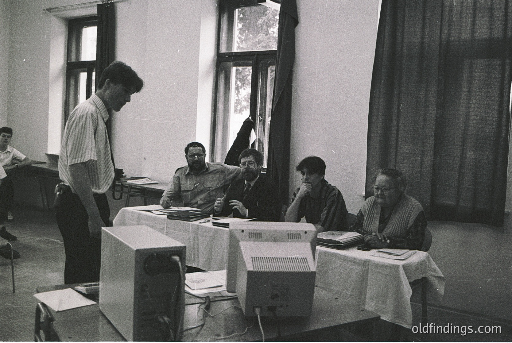 Retro office scene featuring early desktop computers (1980s-era) in a classroom-like setting. Six individuals engage with terminals, likely for training or collaborative work. Cluttered tables, paper notes, and a chalkboard hint at an educational or corporate workshop. --- *Note: If location or specific region is identifiable (e.g., Eastern Bloc due to uniform style), add or similar.*