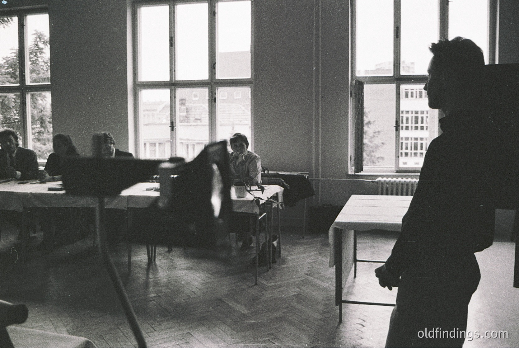 Classroom setting with mid-century industrial design—wooden desks, metal chairs, and large windows revealing urban buildings. Likely Eastern Bloc-era educational institution, 1960s–1980s. Silhouetted figure suggests a teacher or instructor.