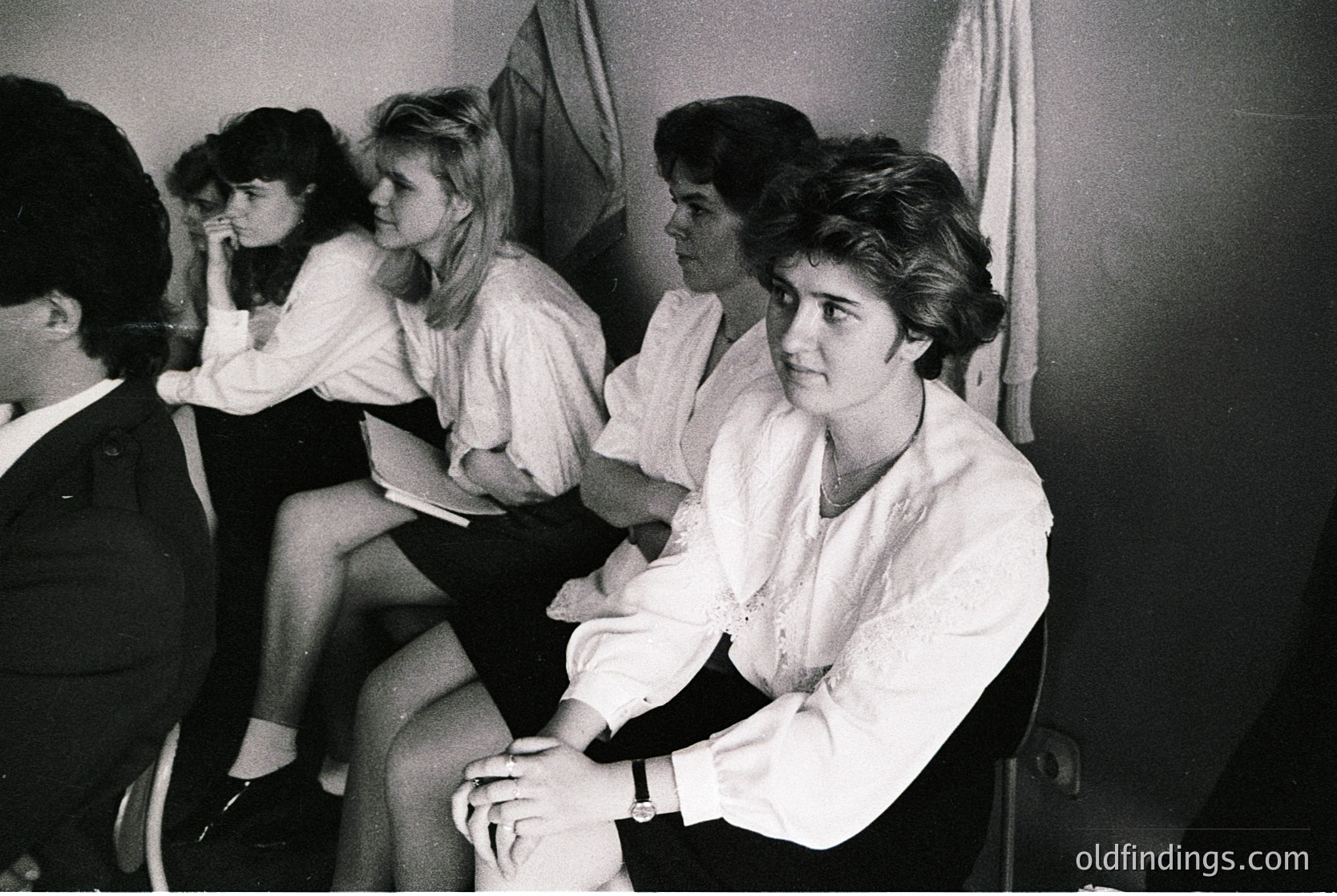 Group of five women in 1970s-era clothing—blouses, knee-length skirts, and short hair—sitting in a dimly lit indoor setting. The central figure wears a white blouse with a watch, while others lean against a curtained backdrop. Likely a social or professional gathering.