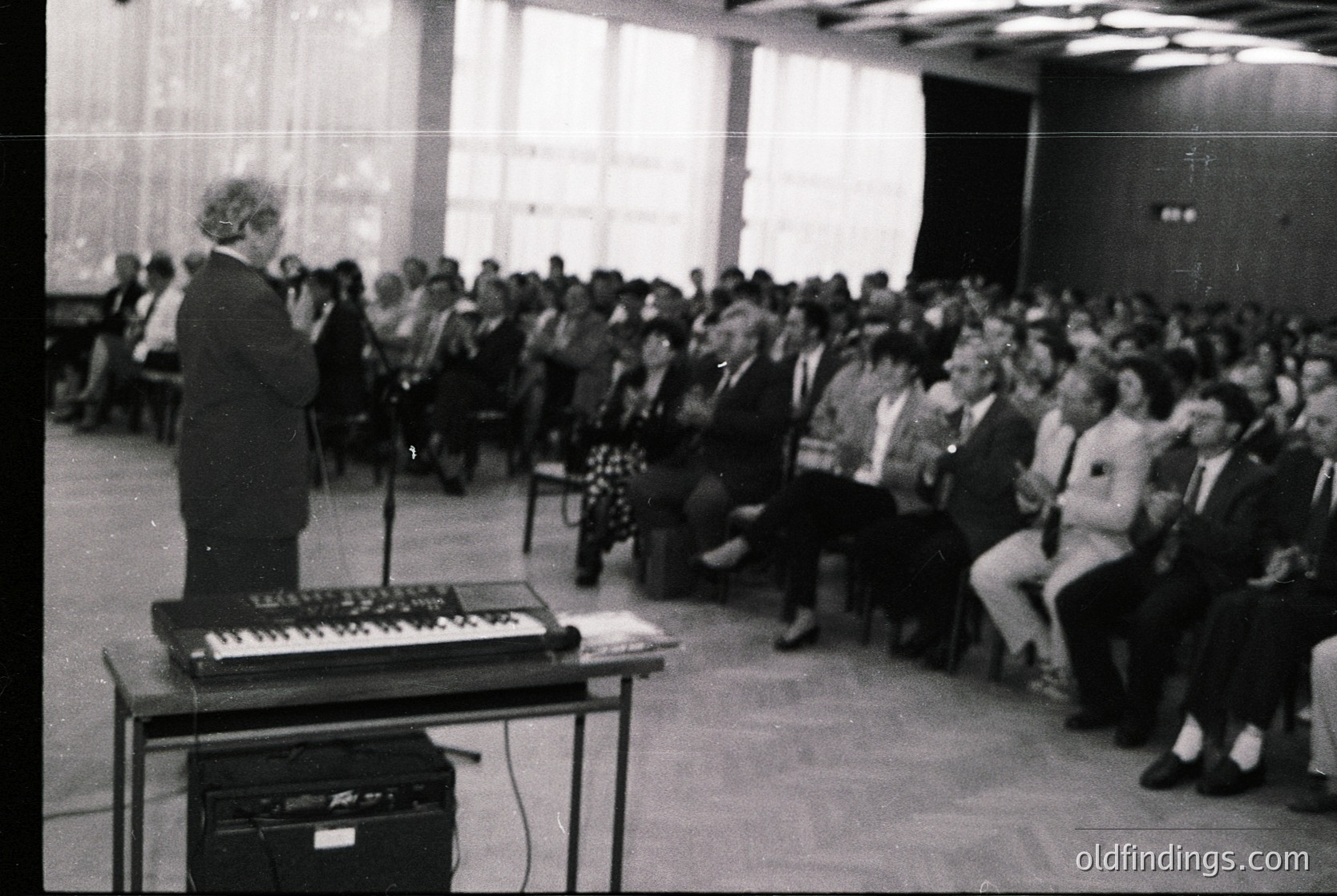 A speaker addresses a packed indoor audience in a formal setting, likely a conference or lecture hall. The speaker stands at a podium with a vintage keyboard instrument, suggesting a 1970s–1980s timeframe. Attendees wear business attire, indicating a professional or academic event. The venue’s minimalist design and lighting reflect mid-century institutional architecture. [Formal lecture hall with vintage keyboard instrument, 1970s–1980s ]