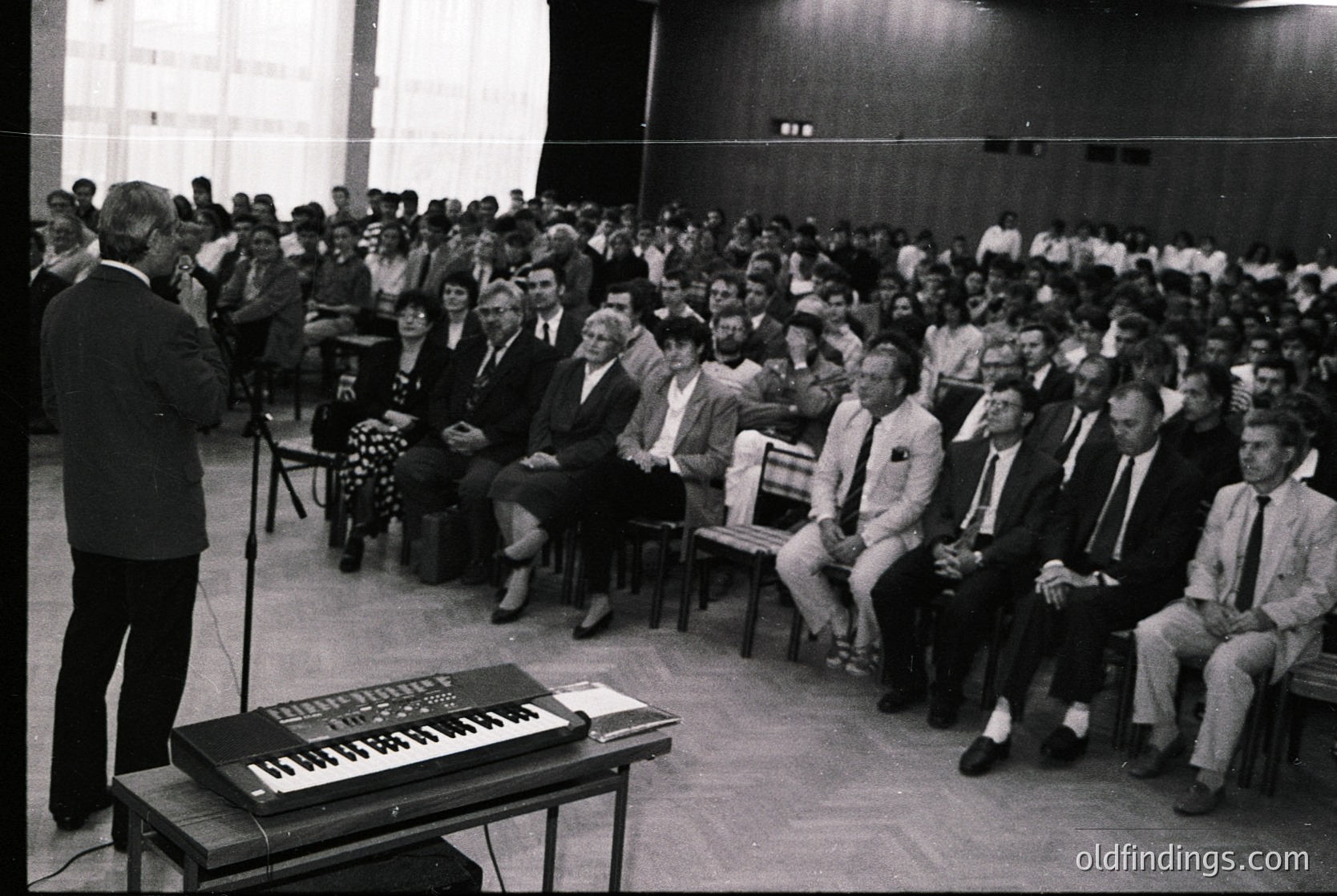Black-and-white lecture hall scene from the 1960s–70s, featuring a speaker at a podium with a keyboard instrument, likely a melodica. Audience of ~150 seated in tiered rows, predominantly men in suits and ties, some women in dresses. Minimalist industrial-style architecture with exposed beams and concrete walls. Potential educational or corporate event setting.