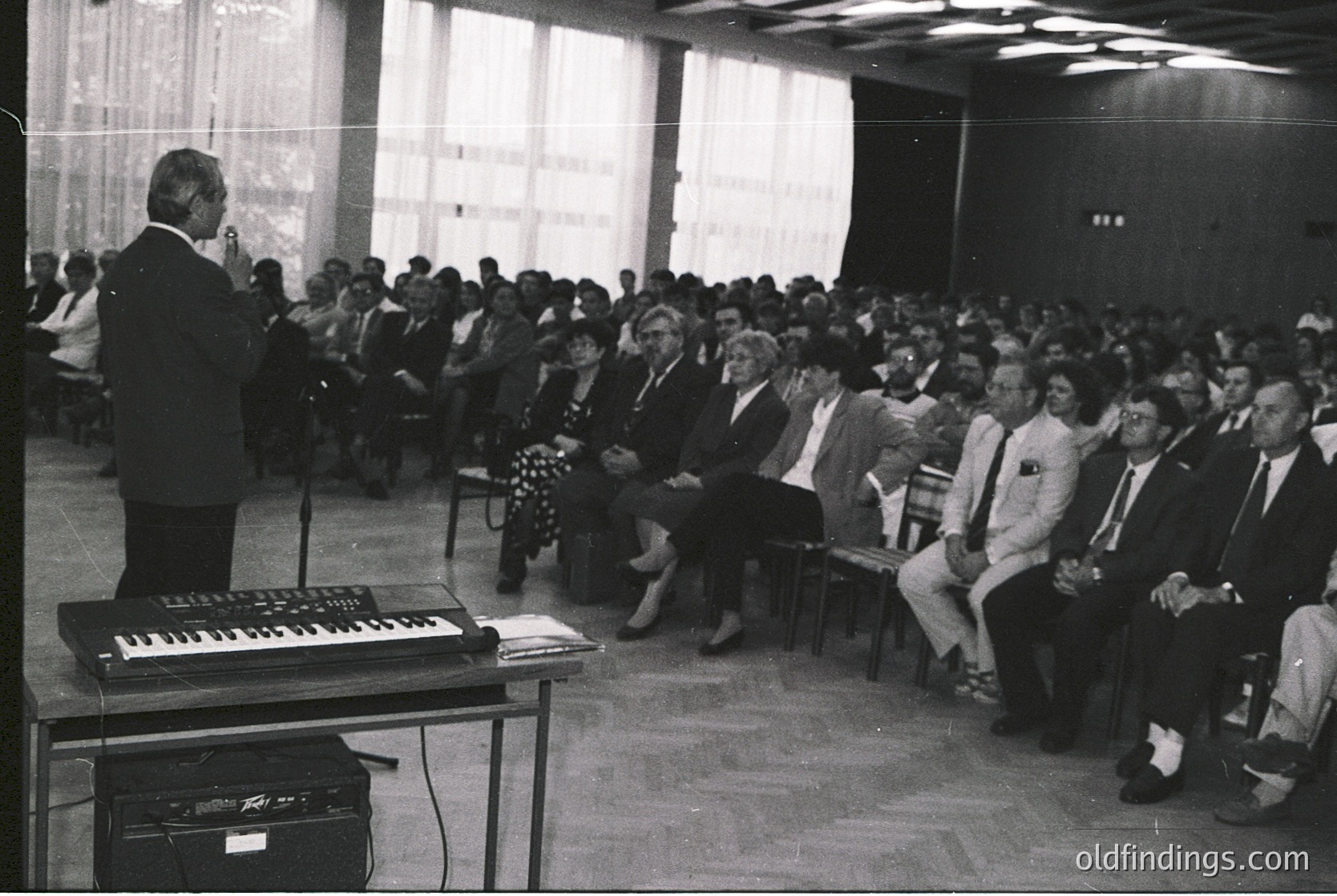 A formal indoor lecture or conference in a mid-20th-century setting, featuring a speaker at a podium with a keyboard instrument (likely a modular synthesizer) and amplifier. Audience seated in rows of wooden chairs, suggesting an institutional or academic venue. Attendees wear suits and ties, indicating a professional or governmental gathering. Natural light filters through sheer curtains.