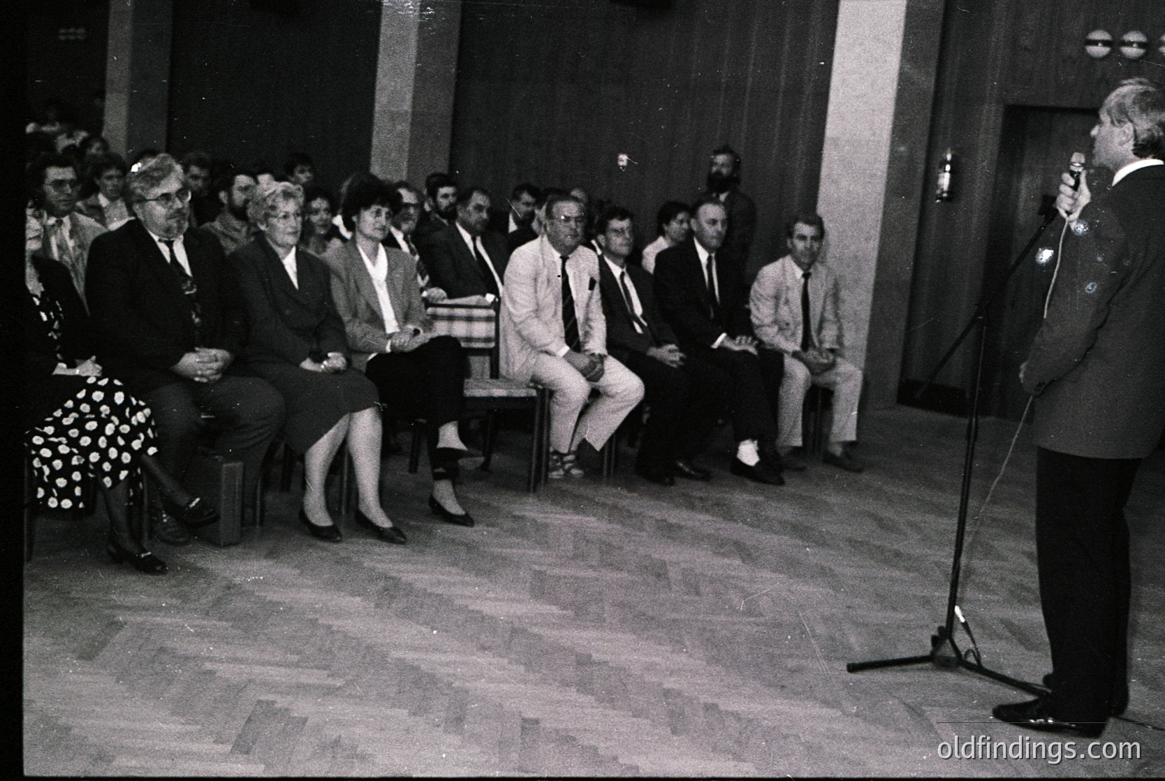 Black-and-white indoor gathering in a formal setting, likely mid-20th century. A speaker stands at a podium with a microphone, addressing an attentive audience seated on wooden benches. Attendees wear business attire: suits, blazers, and dresses. The room features high ceilings, wooden flooring, and a large doorway in the background. Potential event: conference, lecture, or official meeting.