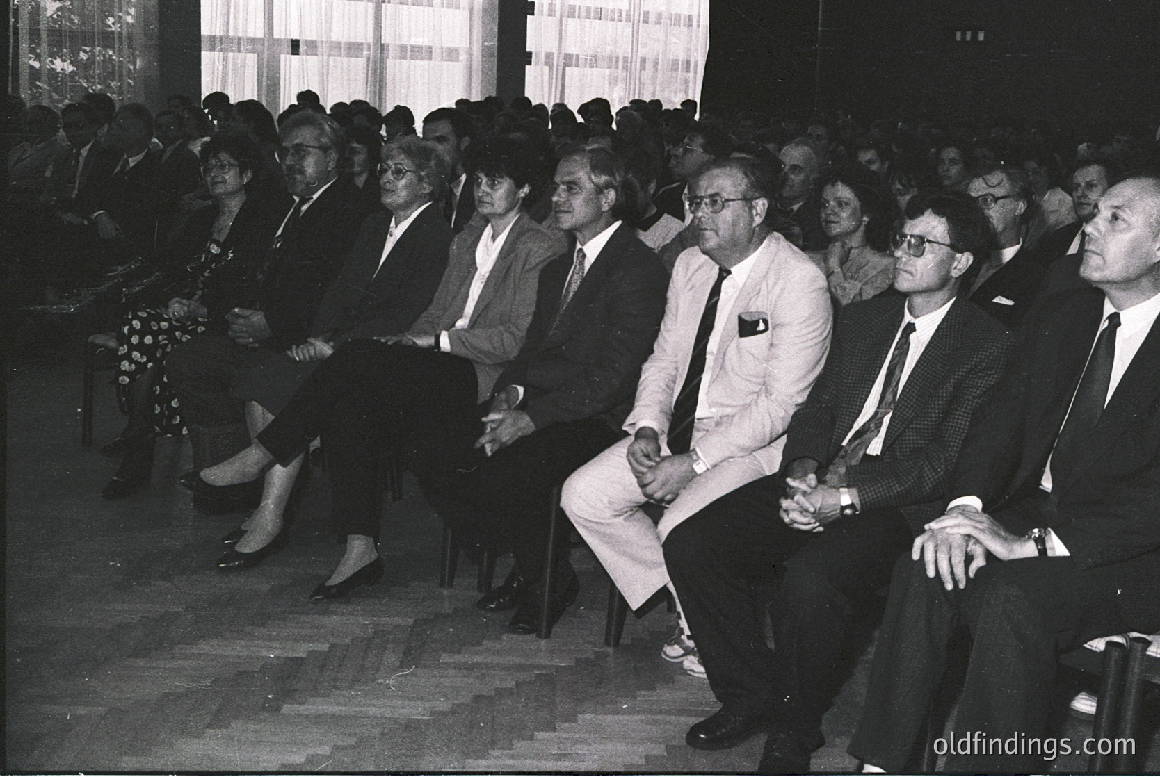 Mid-20th century formal gathering in a tiered auditorium. Attendees in suits, ties, and dresses sit attentively, suggesting a conference or official event. Checkered floor and tiered seating indicate institutional or governmental setting. Natural light from windows highlights formal attire and structured arrangement.
