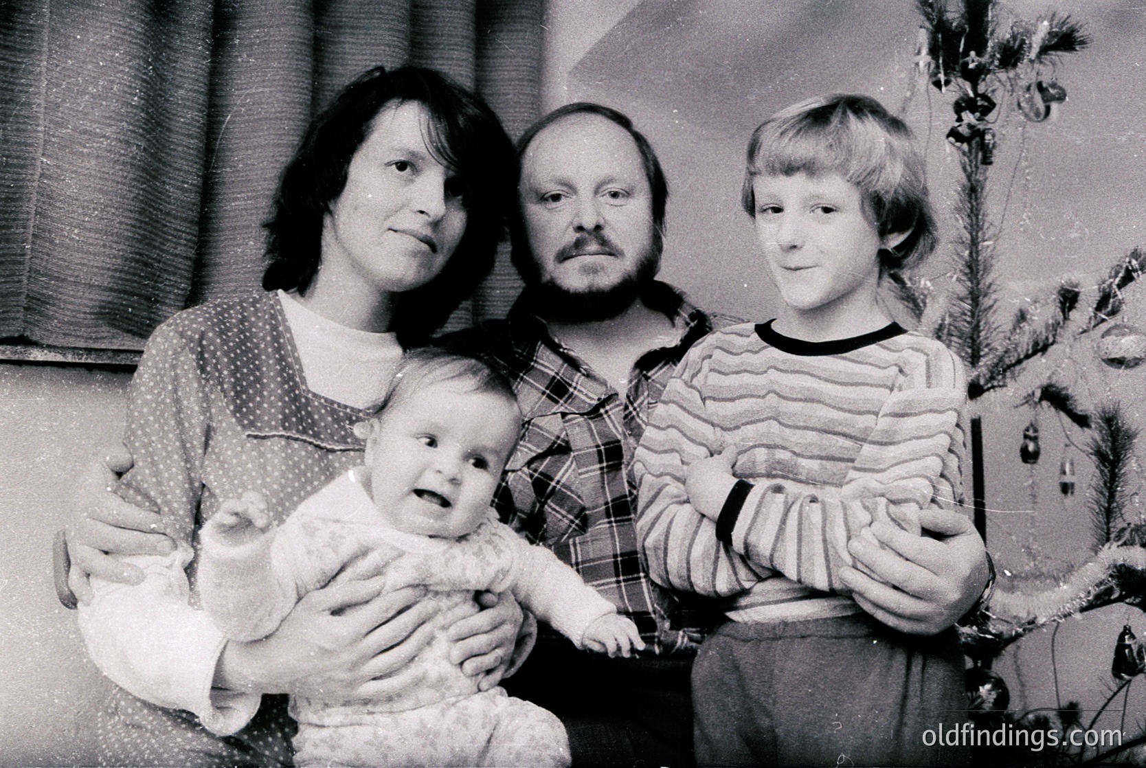 Family portrait from the 1970s–1980s, featuring three adults and an infant. Mother holds baby in a knitted sweater, father wears plaid shirt, son in striped sweater. Indoor setting with Christmas tree and patterned wallpaper.