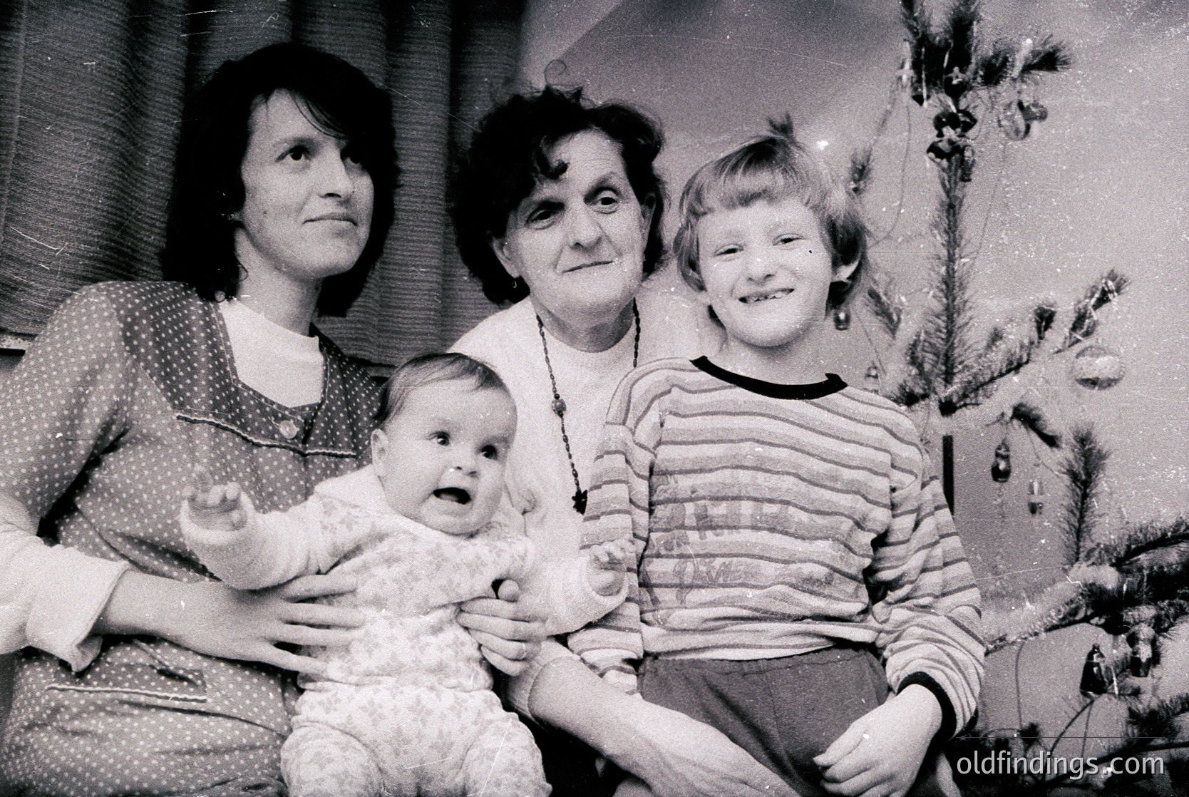 Family portrait from the 1960s–1970s, featuring three generations: a woman holding a baby, a seated elderly woman, and a young girl. Indoor setting with a decorated Christmas tree in the background. Mid-century clothing and hairstyles suggest Western European or North American context.