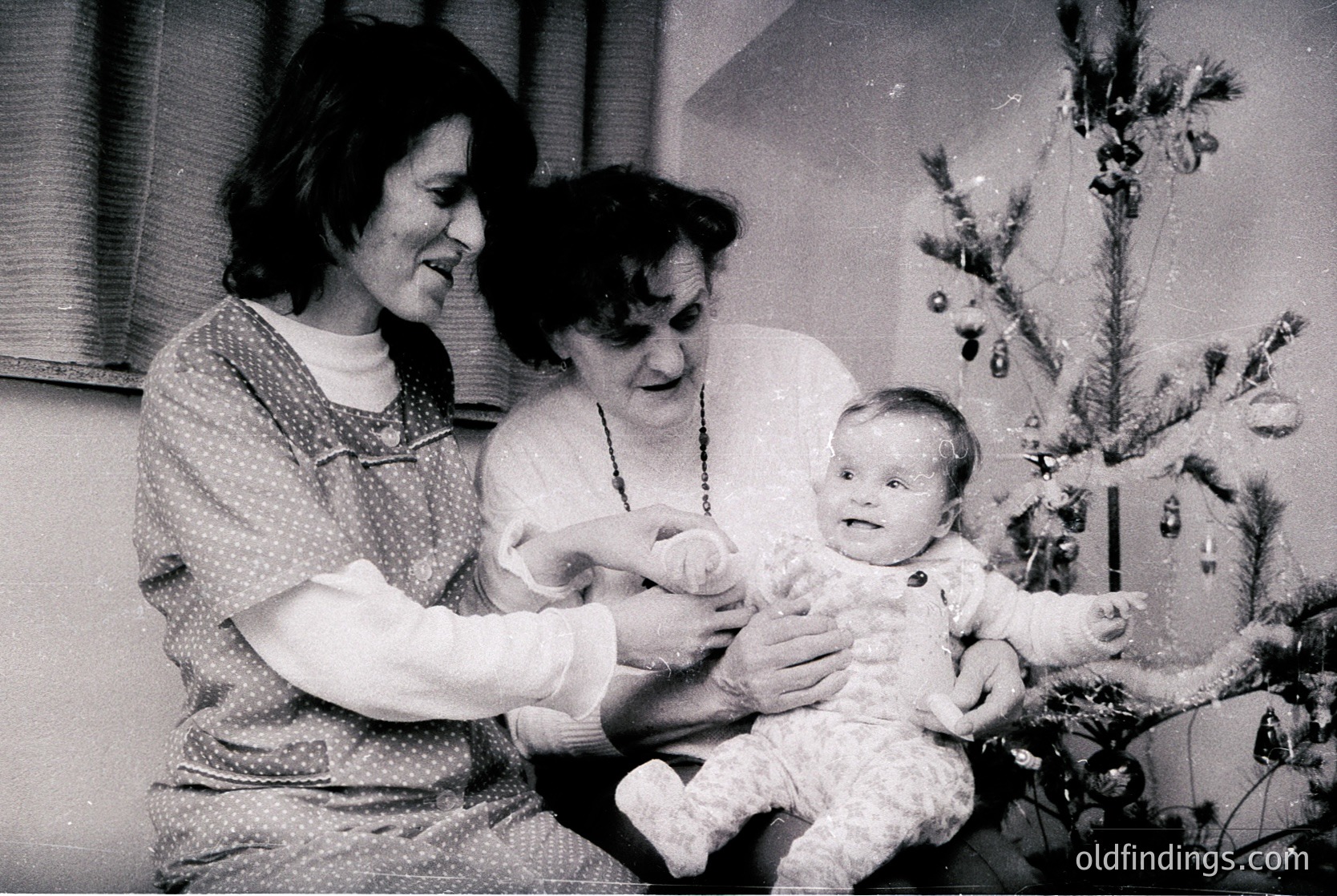 Two women cradle a baby in a festive indoor setting, likely a 1960s-1970s Christmas scene. The woman on the left wears a patterned blouse; the older woman on the right holds a pearl necklace. A decorated Christmas tree with ornaments and lights fills the background.