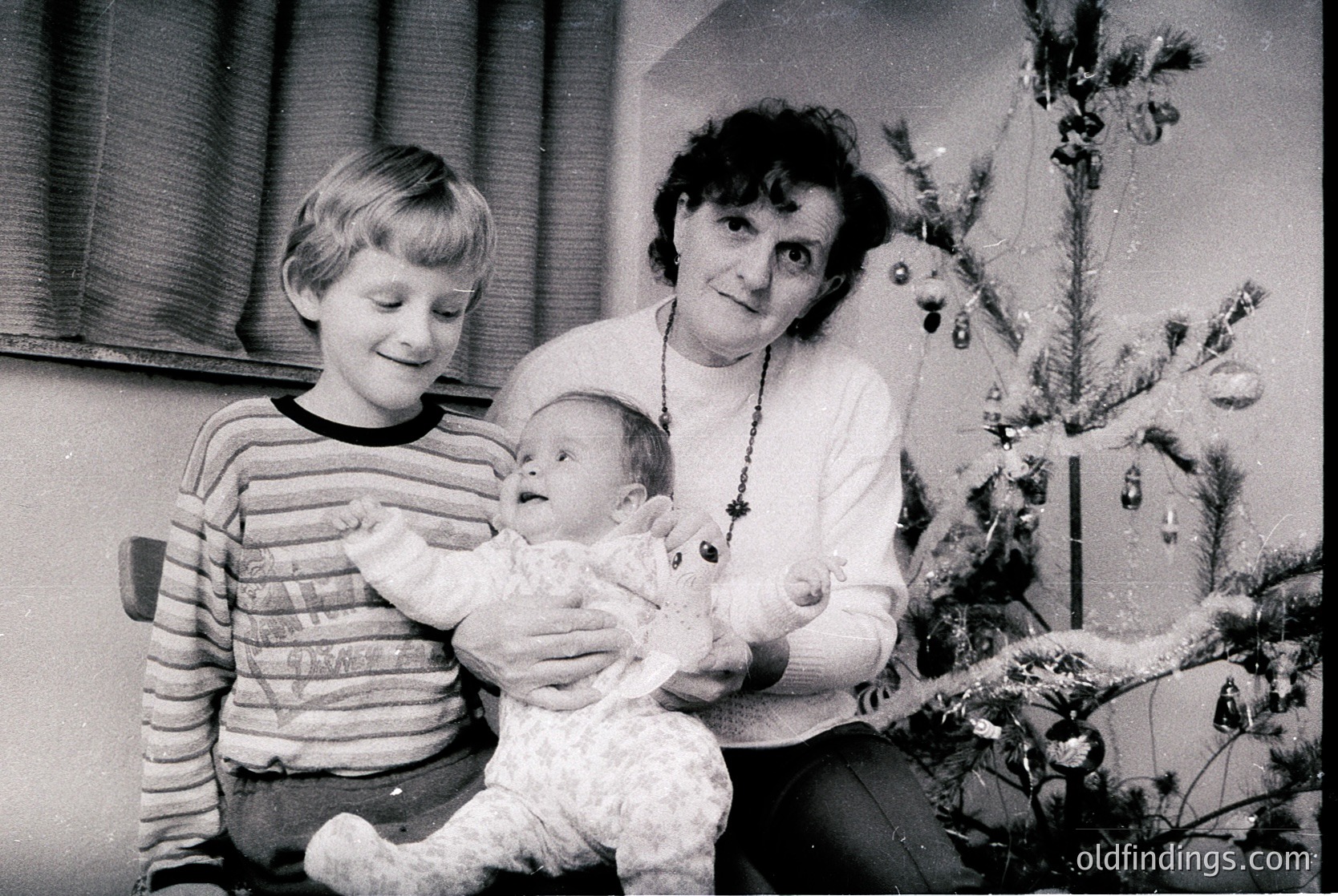A mid-20th-century (likely 1960s–1970s) black-and-white family portrait: a woman cradles a baby in a plush teddy bear outfit, while a young boy stands beside her. Decorated Christmas tree with ornaments and tinsel in background. Warm, intimate holiday scene.