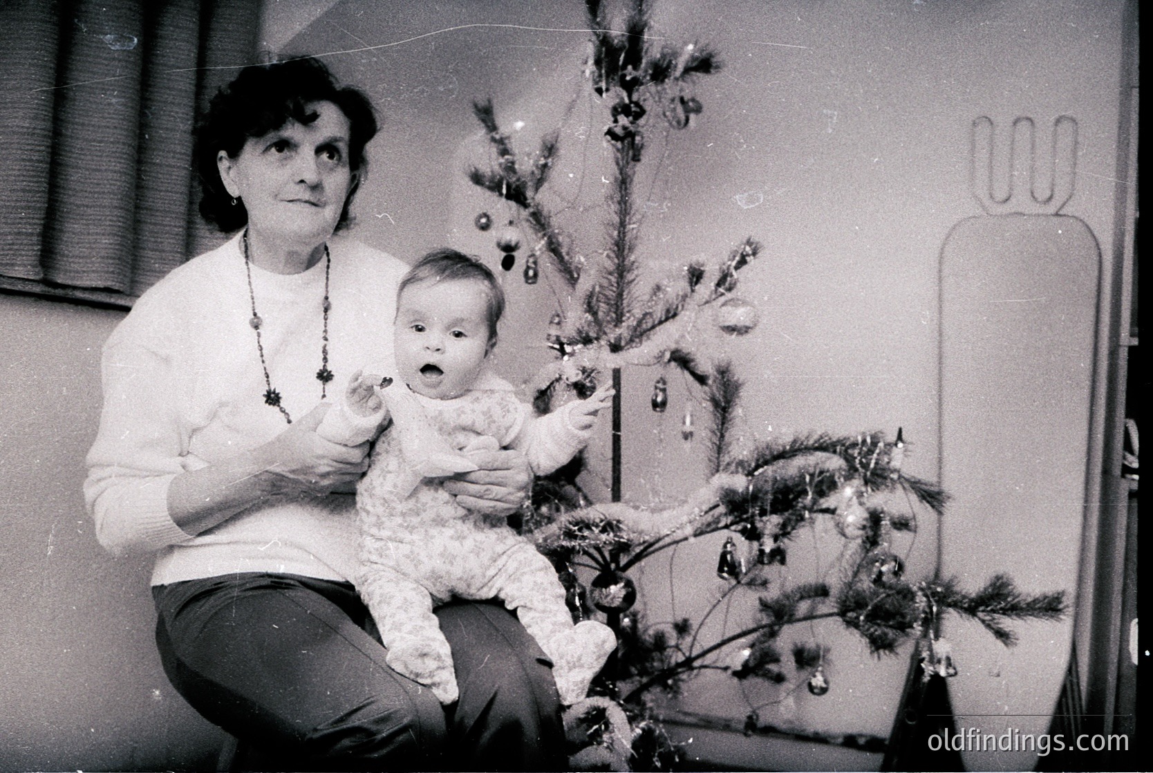A black-and-white snapshot from the mid-20th century (likely 1950s–1970s) captures a tender holiday moment. An adult holds a baby in a cozy sweater beside a modestly decorated Christmas tree adorned with ornaments and tinsel. The setting appears to be an indoor living space with a vintage radiator and simple furnishings. [Mid-20th-century holiday portrait ]